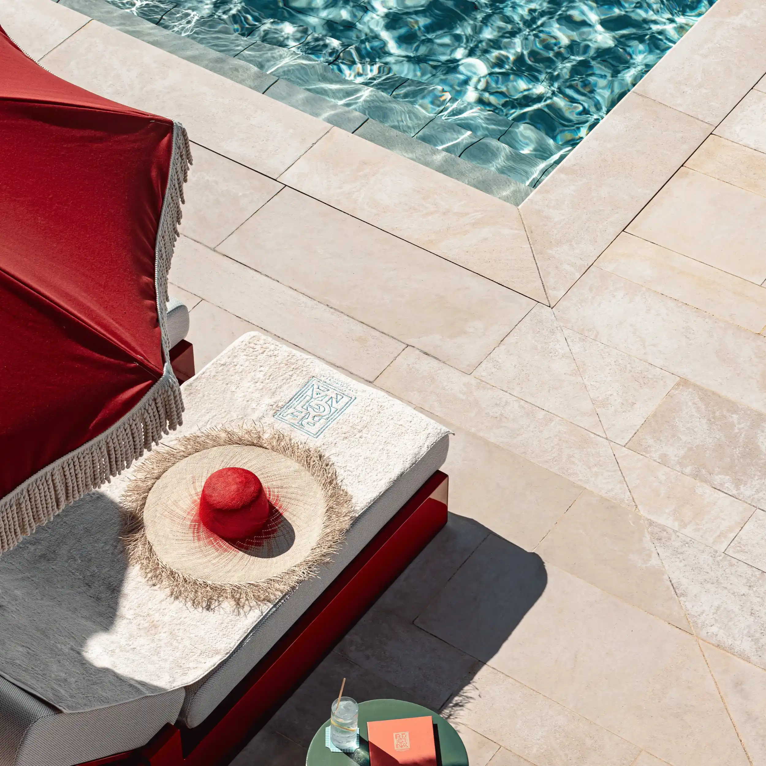 Straw sun hat with a red center on a lounge chair near a pool and a small table with a drink.