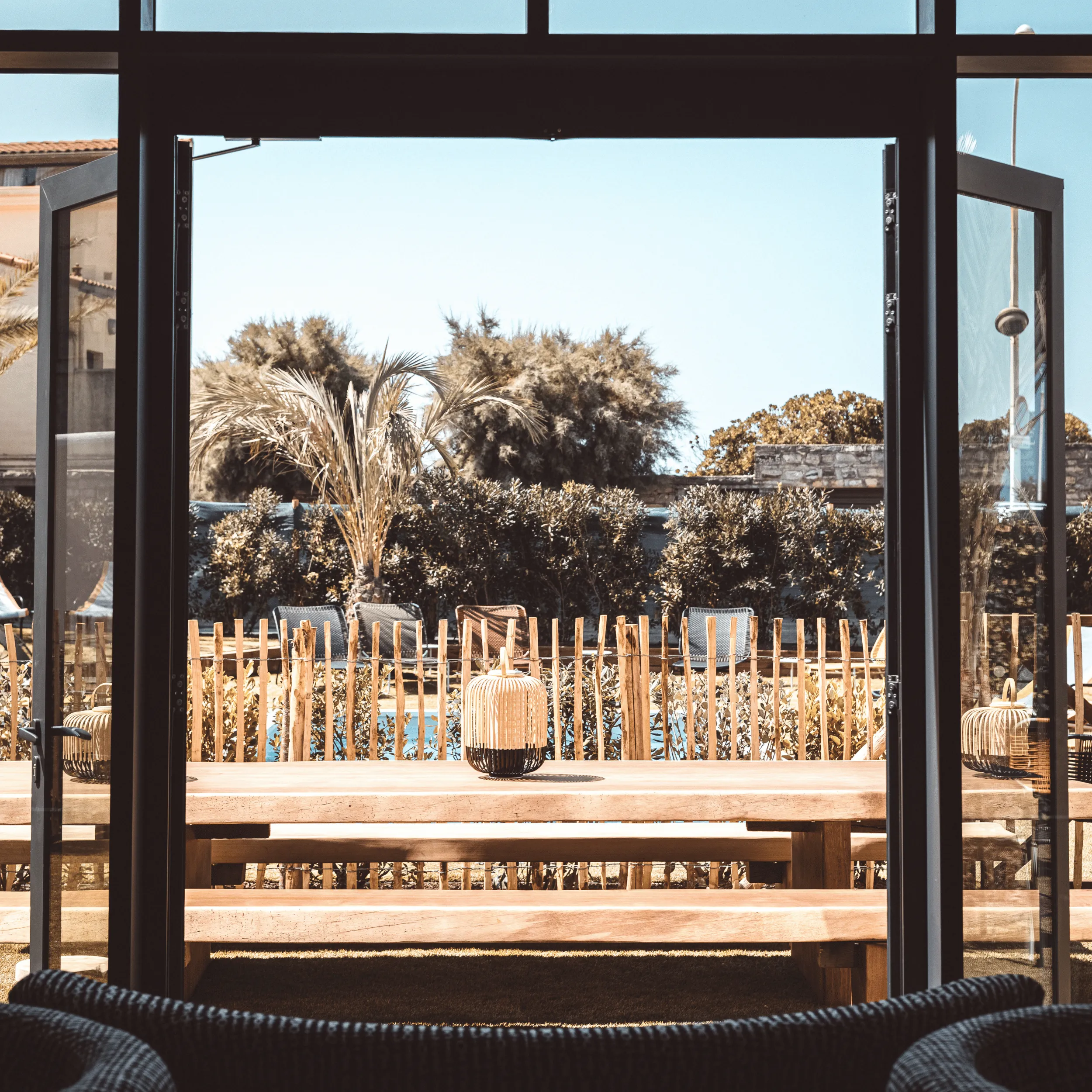Outdoor wooden table and benches seen through open glass doors with plants and blue sky beyond.