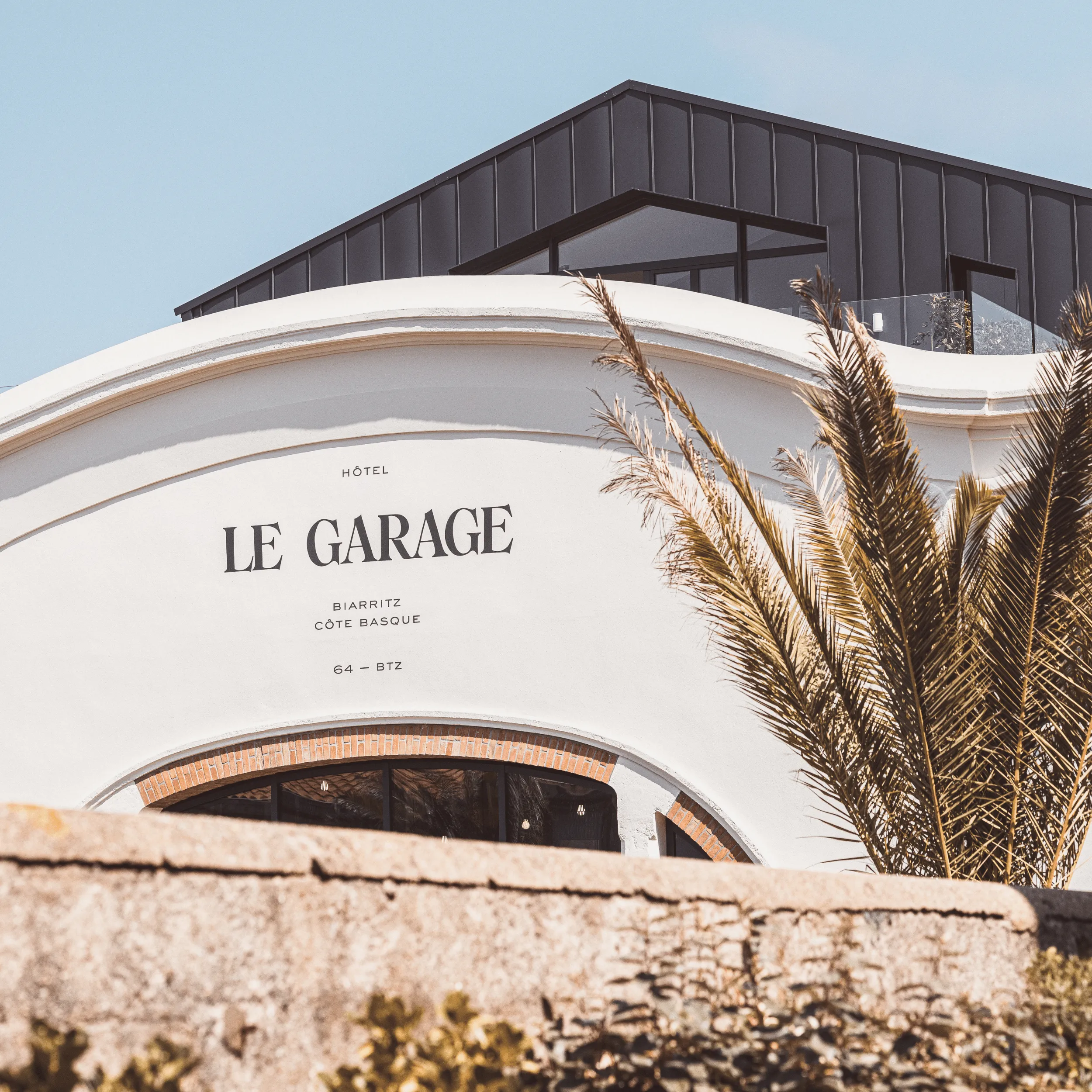 White curved building facade with Le Garage hotel sign in Biarritz, palm tree, modern dark roof above.