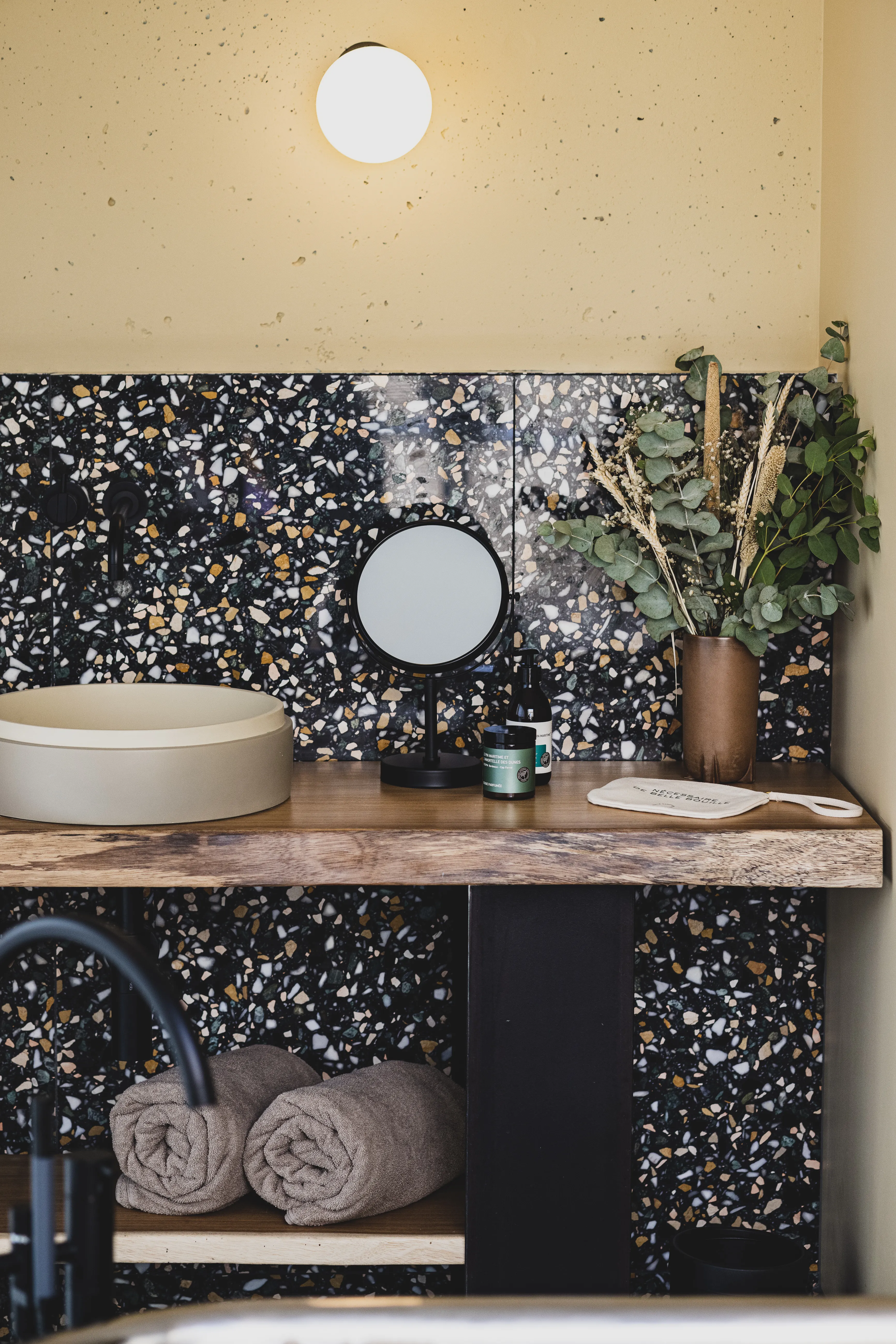 Modern bathroom with terrazzo wall, wooden countertop, round sink, mirror, and decorative plant.