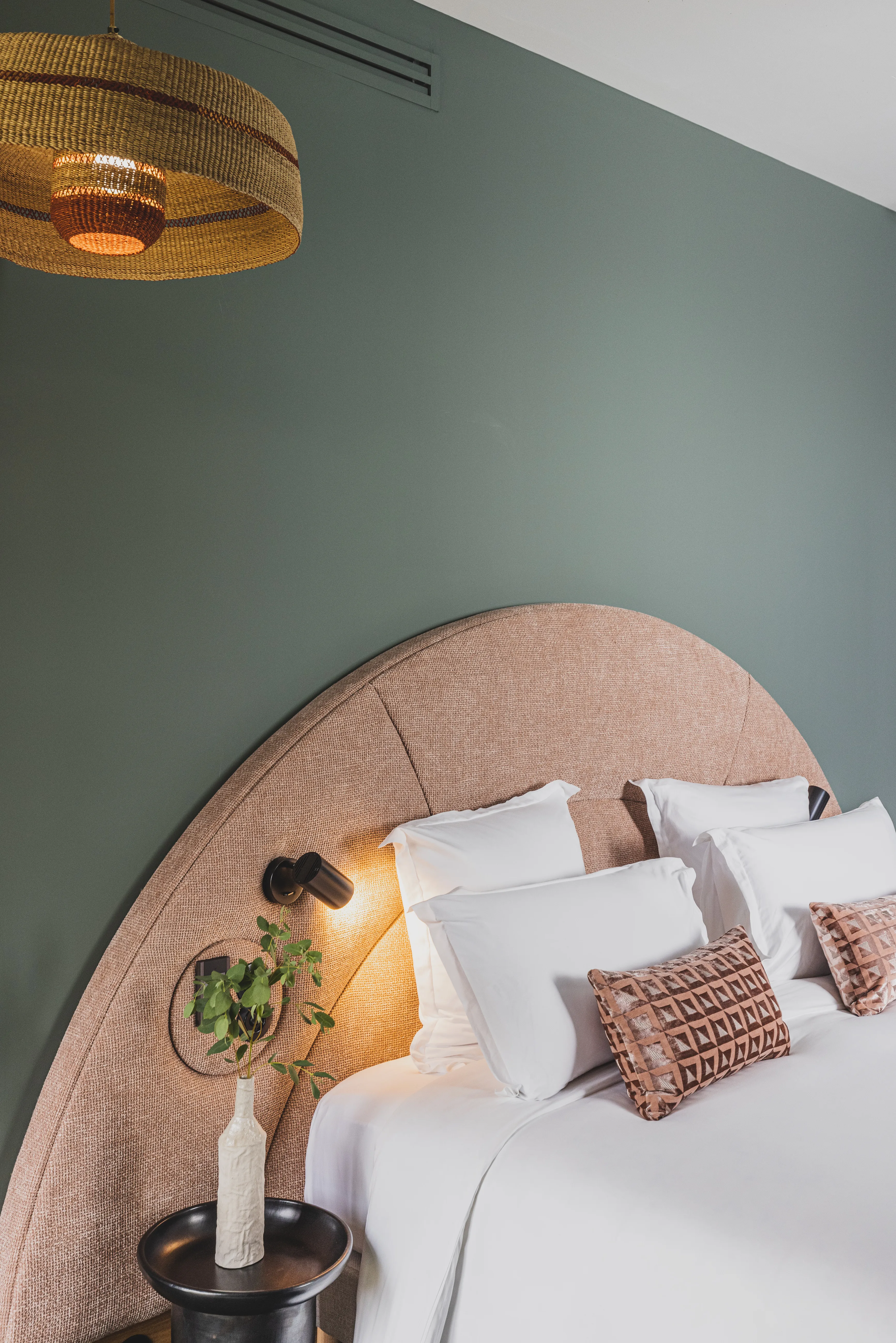 Bed with a curved beige headboard, white pillows, patterned cushions, and a bedside table with a vase.