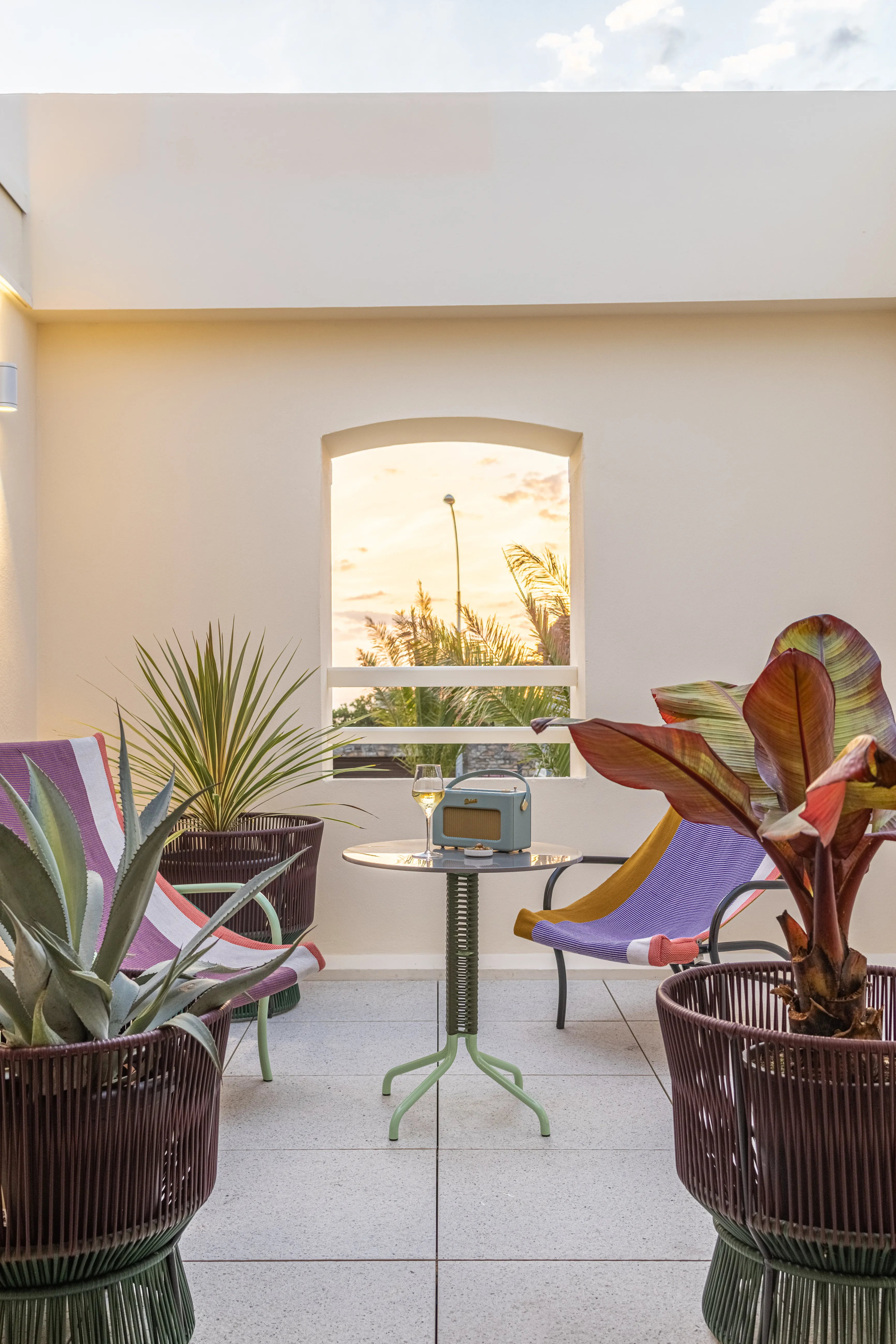 Cozy outdoor patio with colorful chairs, potted plants, a small table, radio, and glass of wine at sunset.