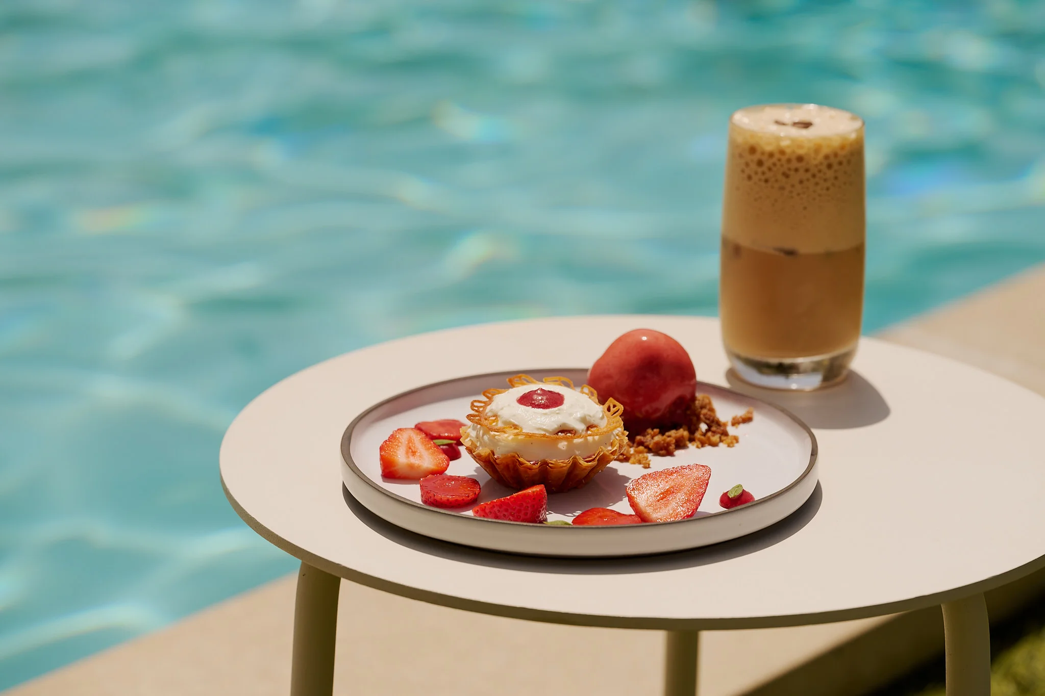 Dessert plate with strawberries, cream tart, fruit sorbet, and iced coffee by a pool on a round table.