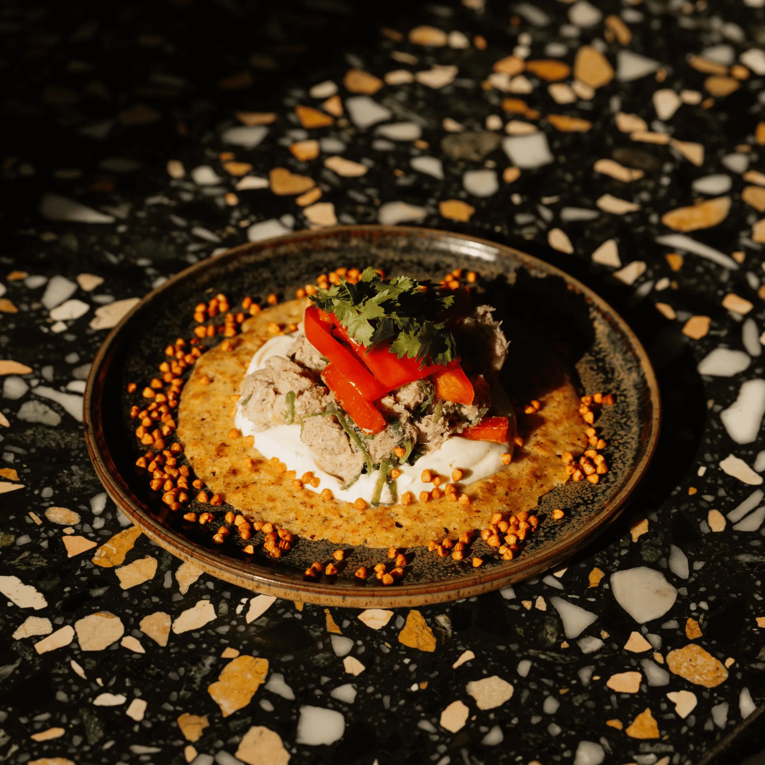 Gourmet dish on dark plate with creamy base, tuna, red pepper slices, herbs, and orange crunchy garnish.