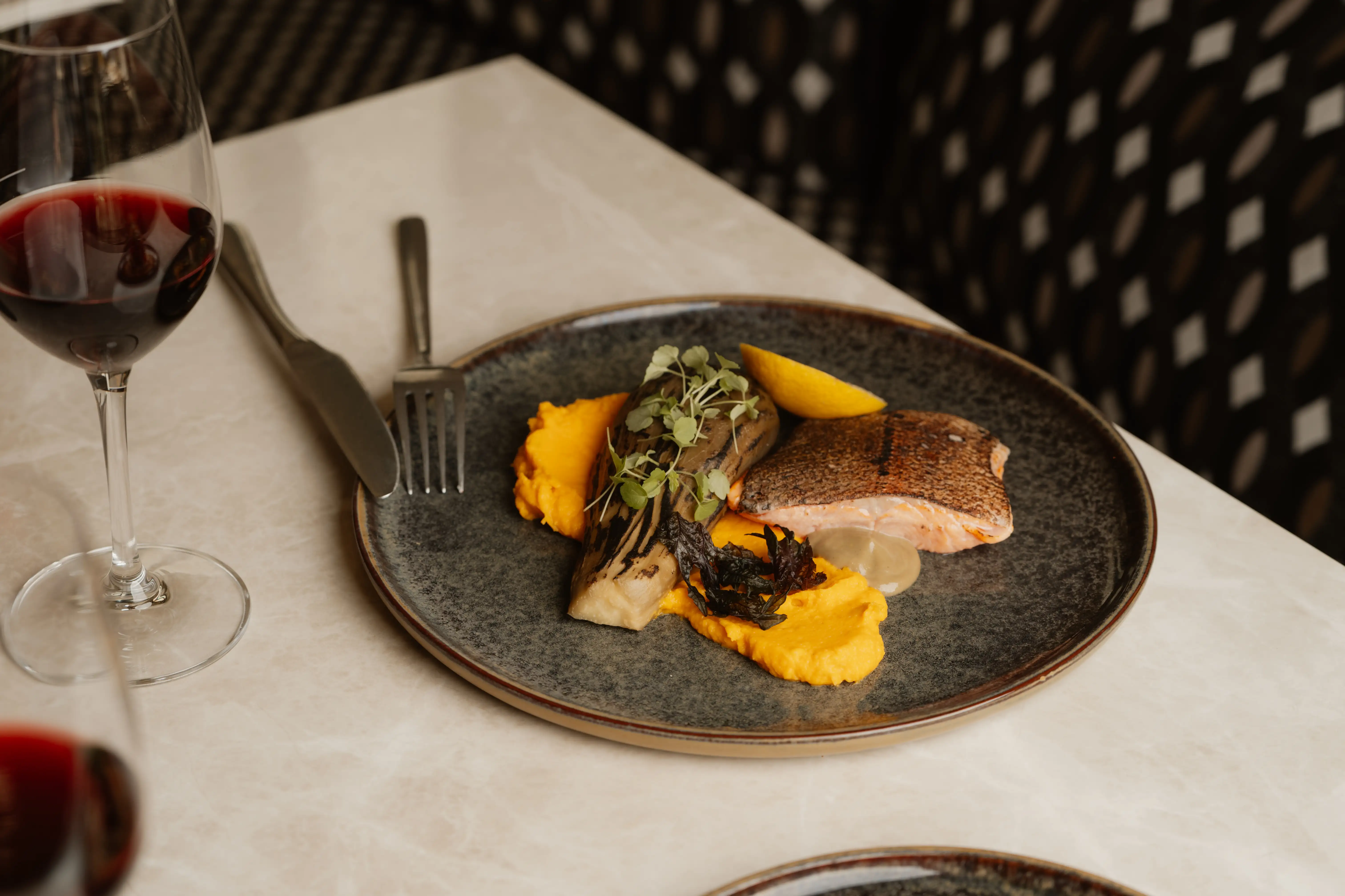 Dish with grilled fish, mashed orange vegetables, eggplant, microgreens, and a lemon wedge next to a glass of red wine.