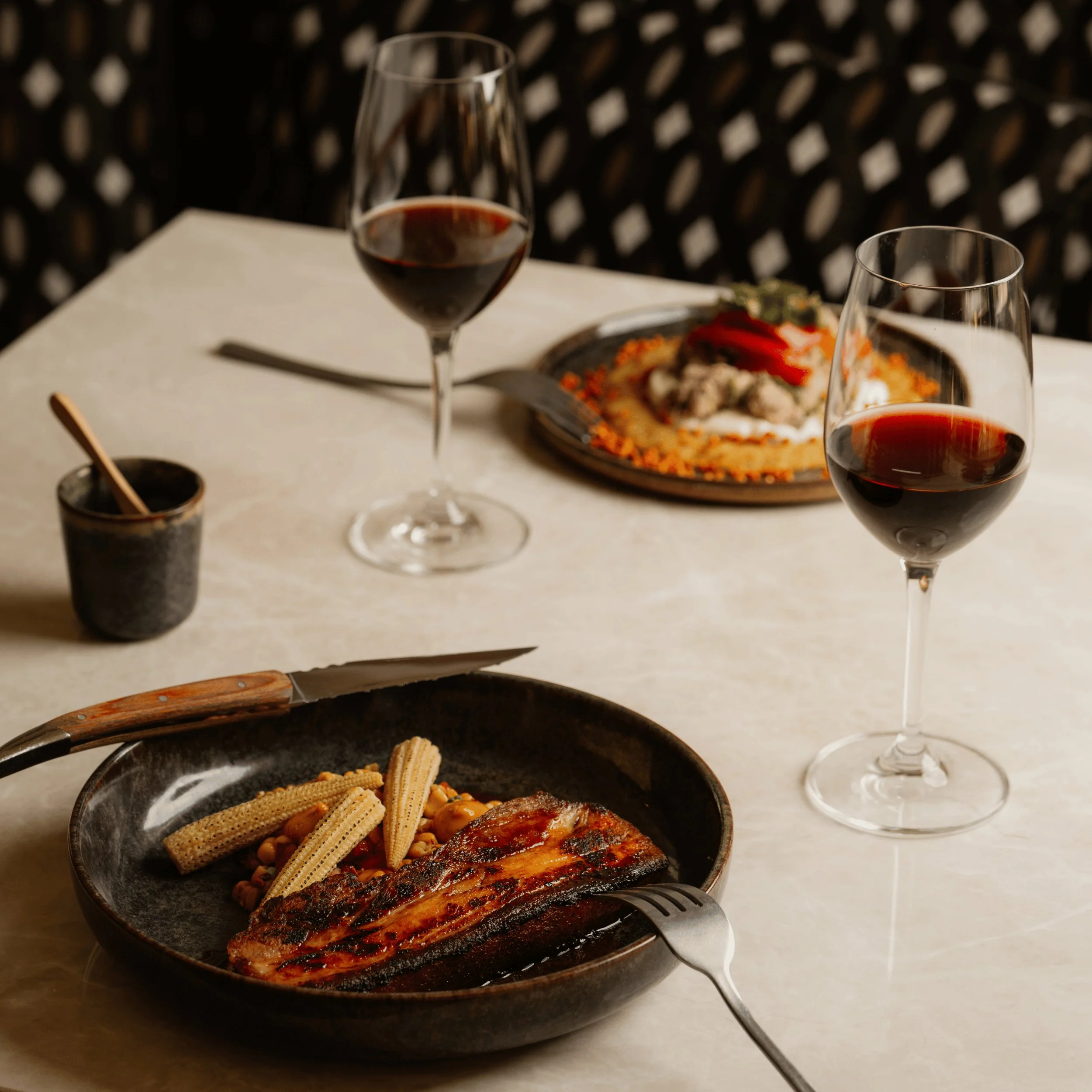 Two plates with gourmet dishes, each paired with a glass of red wine on a marble table.