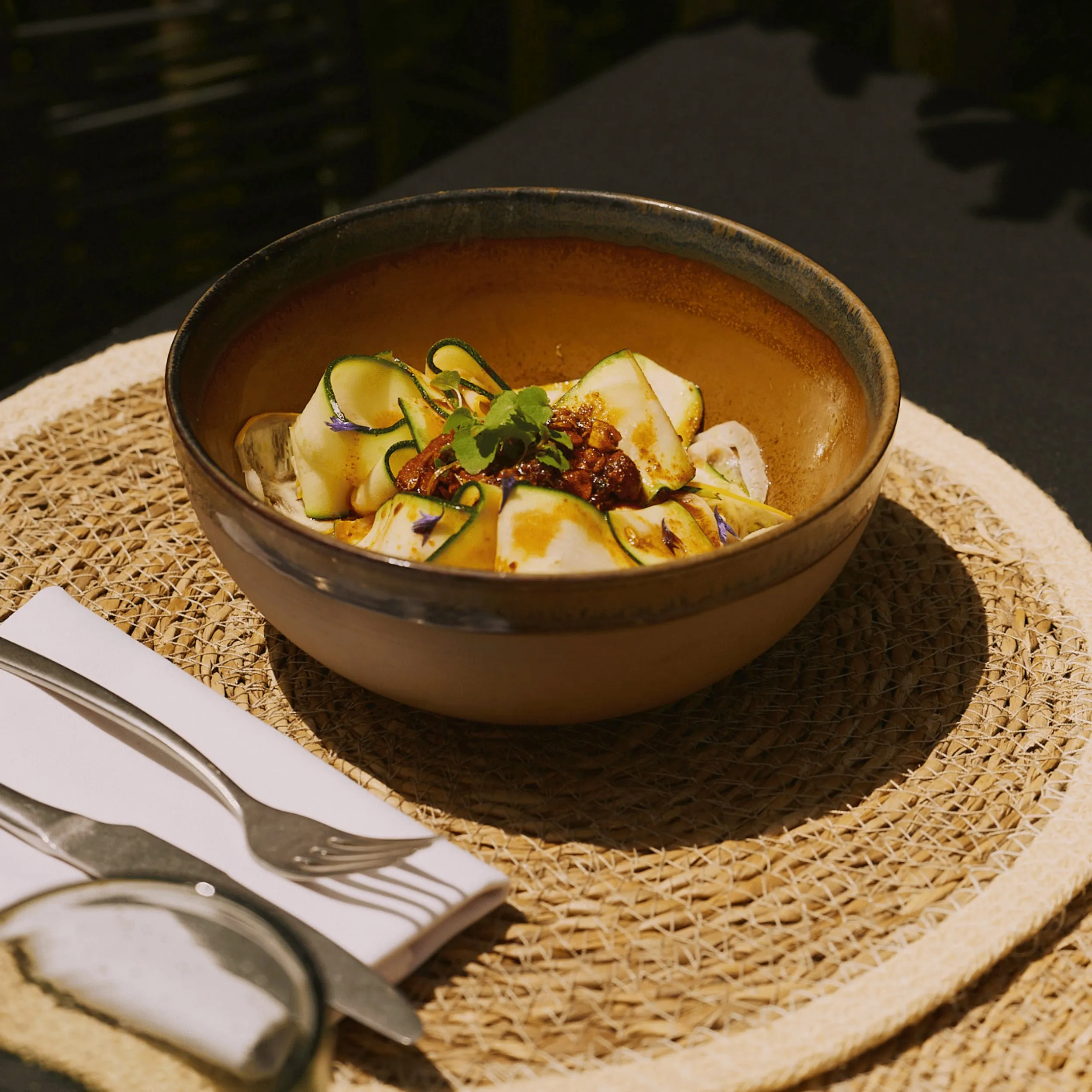 Brown bowl with thinly sliced zucchini, herbs, and sauce on a woven placemat next to cutlery.
