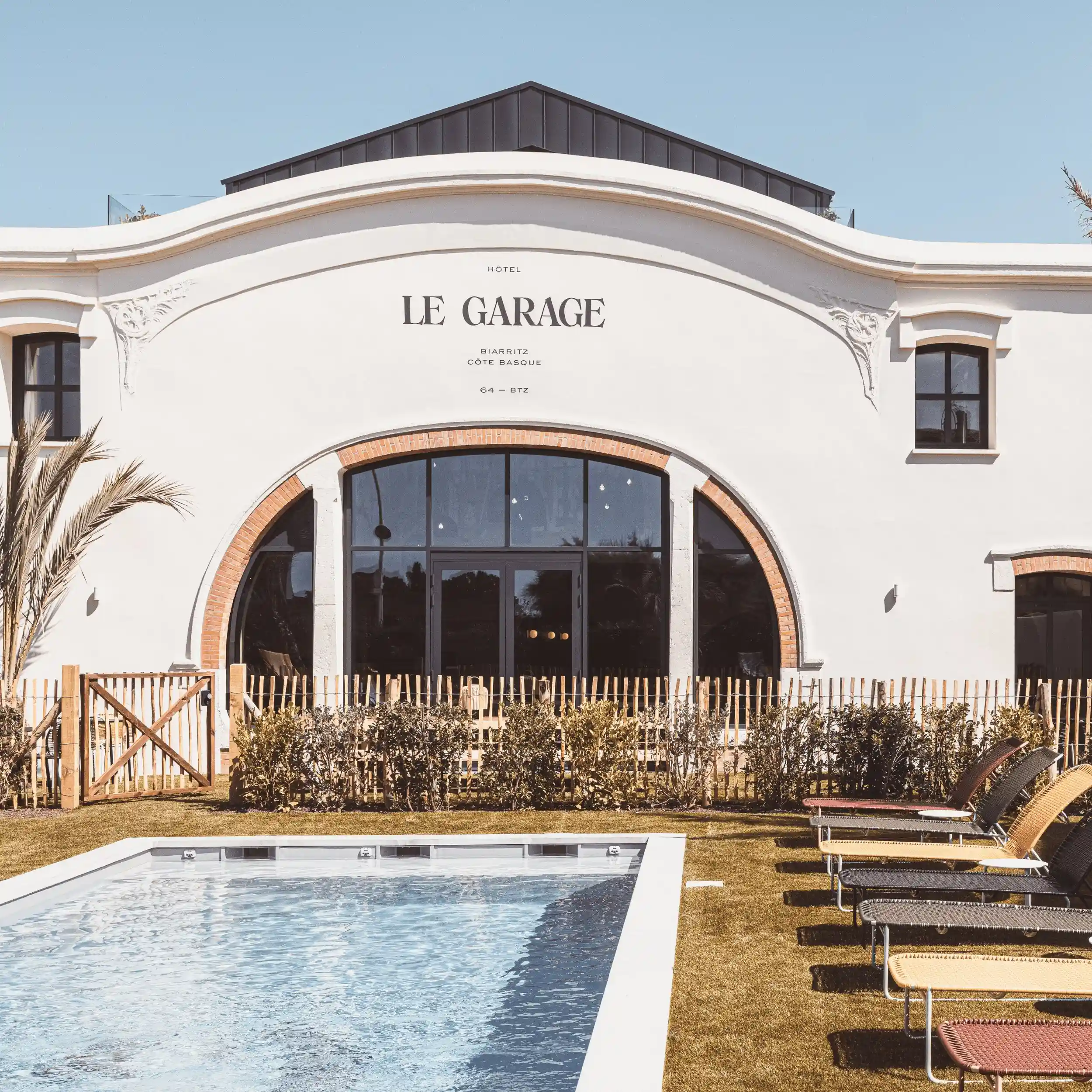 Outdoor pool and lounge chairs in front of Le Garage hotel with a white facade and large arched window.