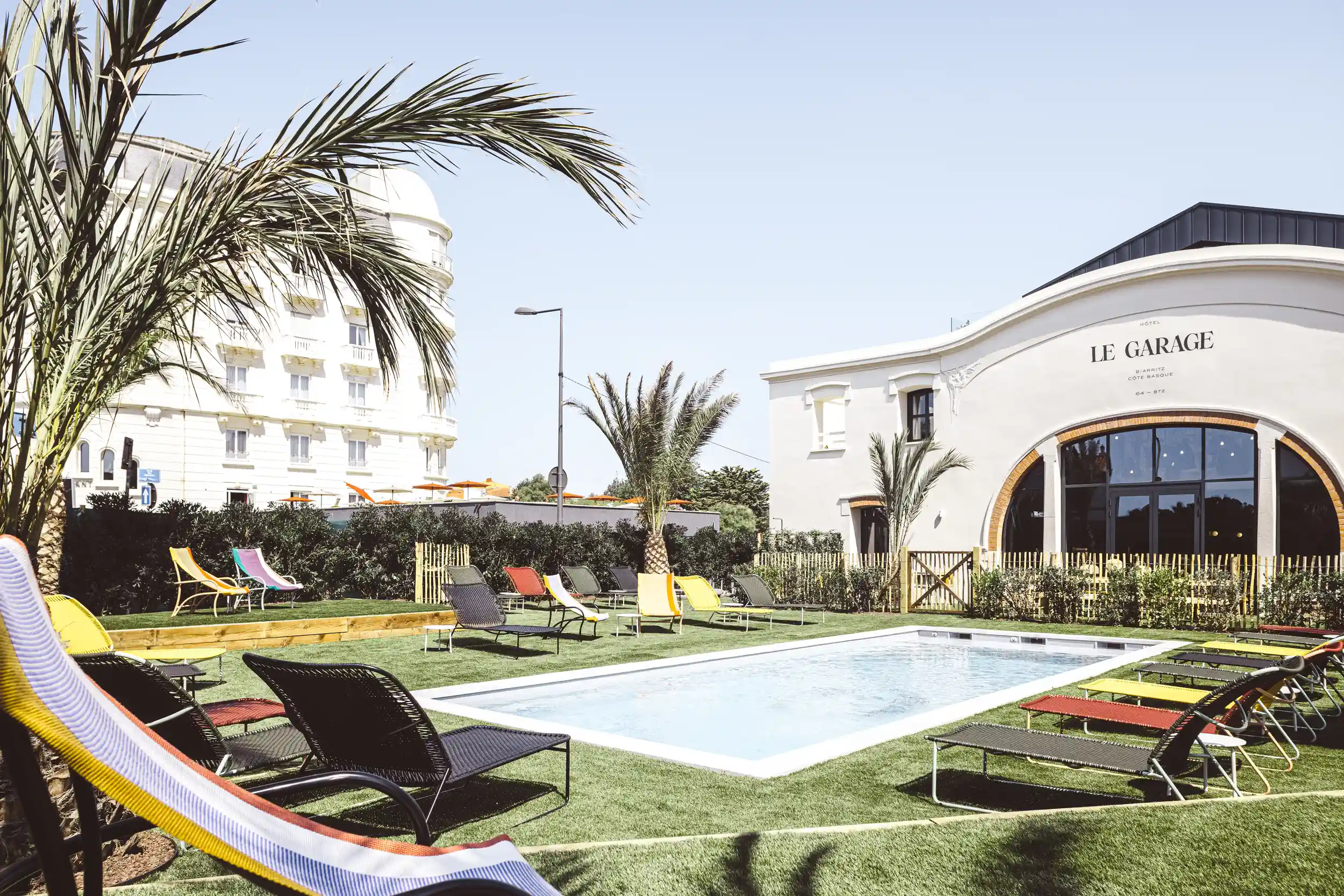 Outdoor pool with colorful lounge chairs on artificial grass near Le Garage hotel building and palm trees.