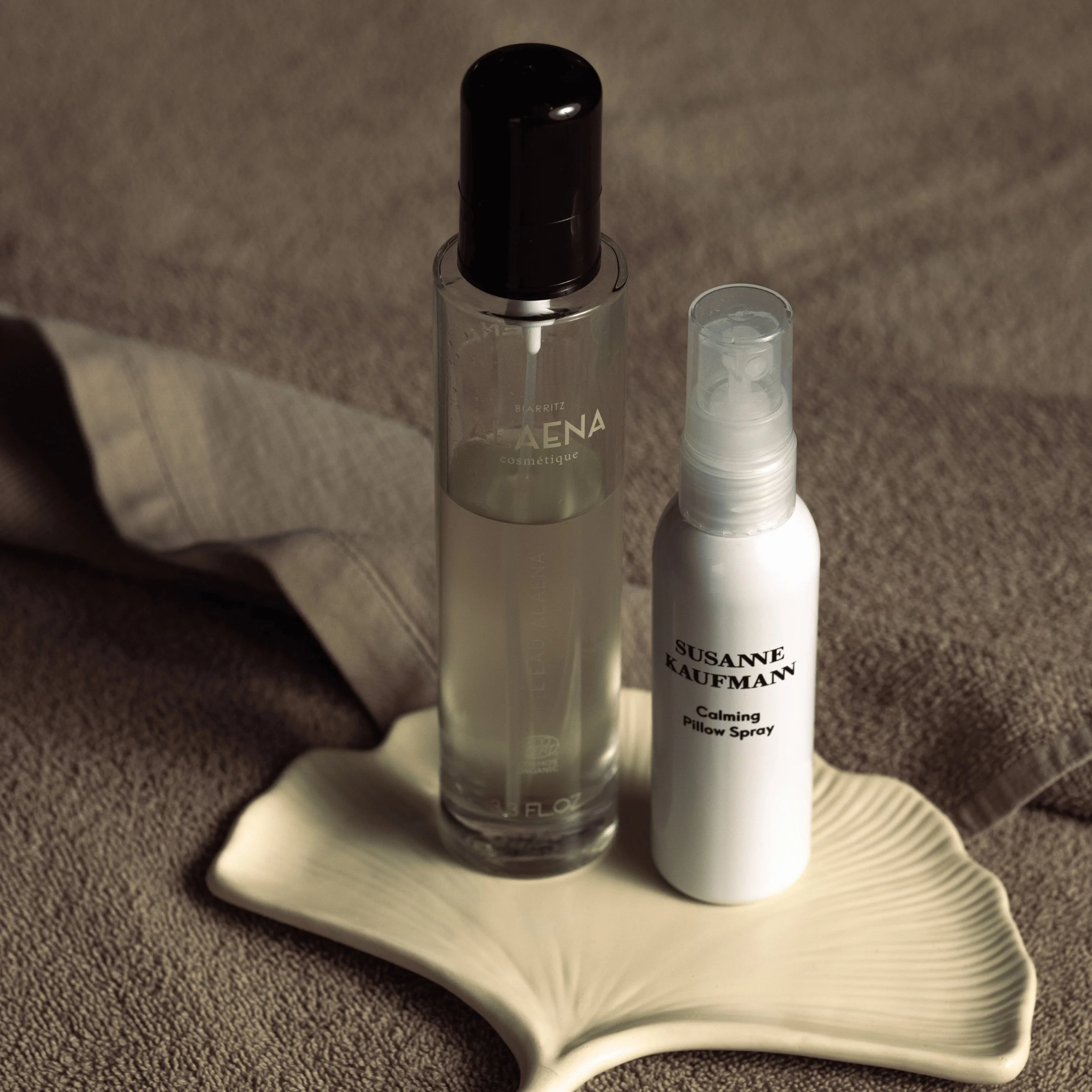 Two cosmetic spray bottles placed on a beige ceramic leaf dish over a soft brown fabric surface.