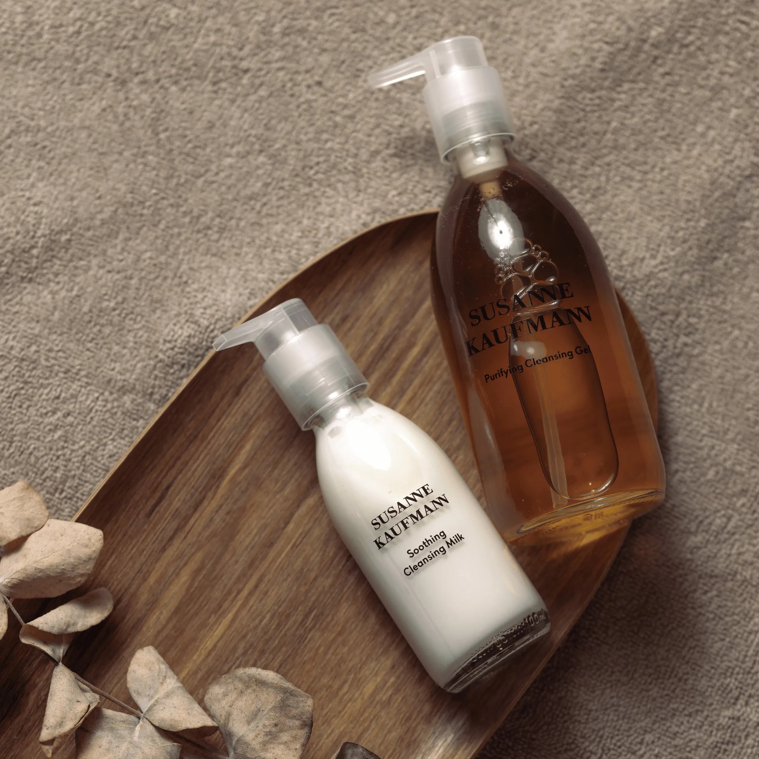 Two Susanne Kaufmann skincare bottles, one white Soothing Cleansing Milk and one amber Purifying Cleansing Gel.