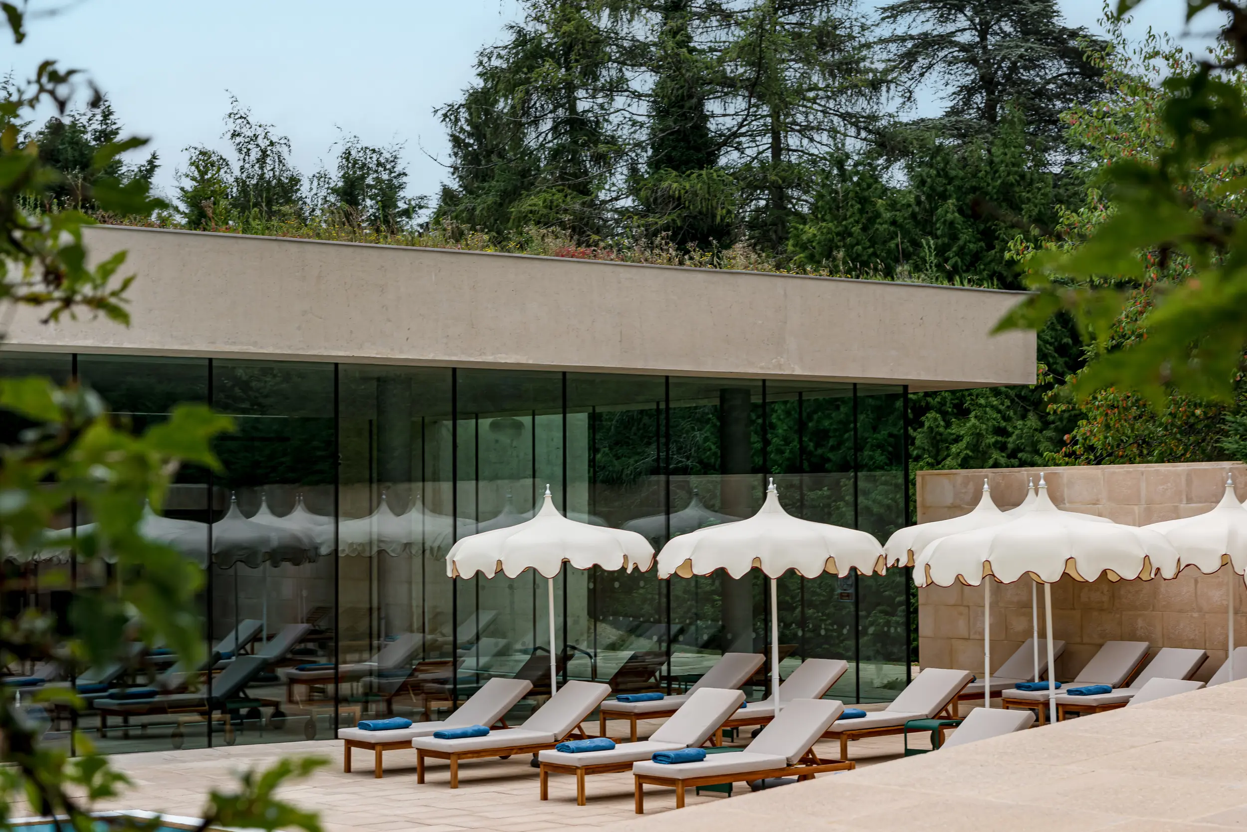 Outdoor lounge area with white umbrellas and cushioned sunbeds beside glass-walled building and trees.