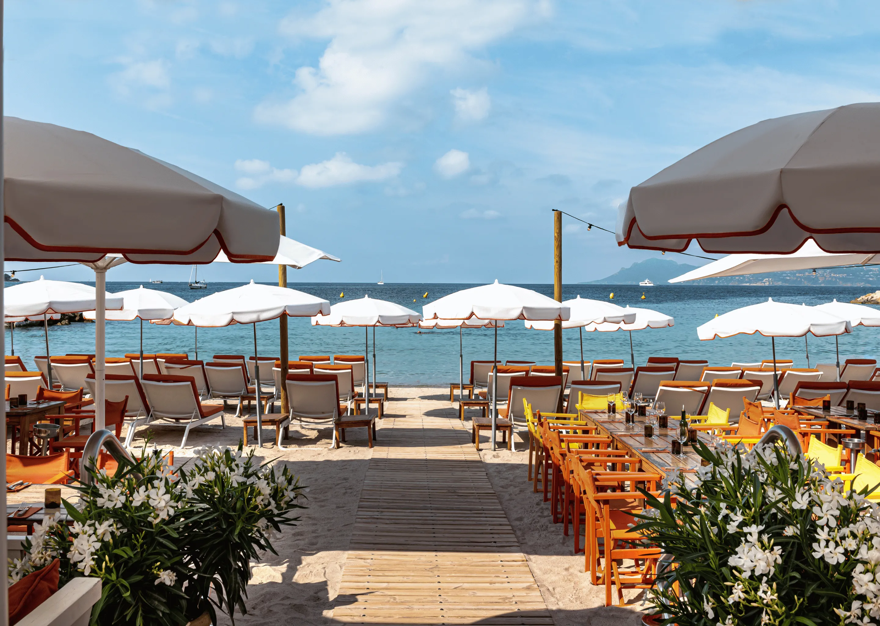 Wooden walkway leads to beach chairs and white umbrellas by the sea with boats and island in background.