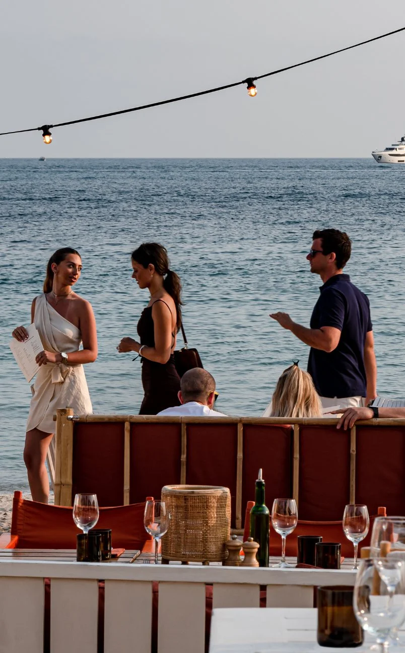 People socializing by the beach with tables and lit string lights at sunset.