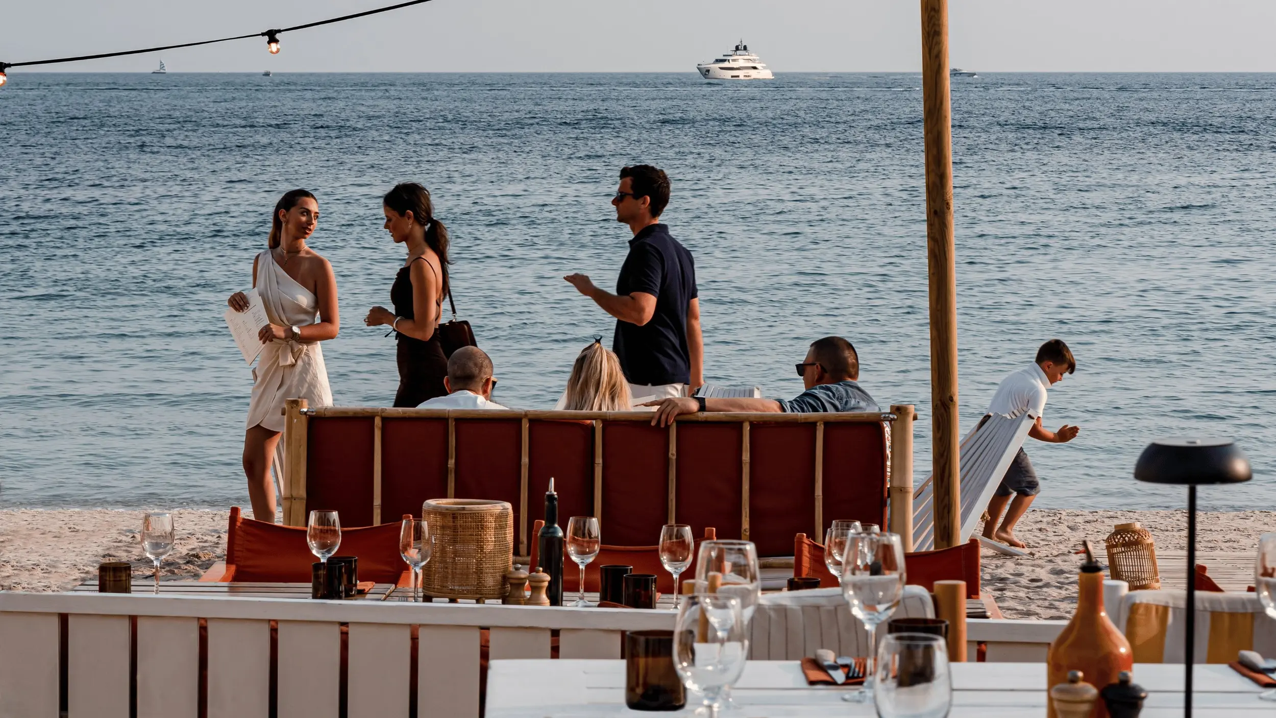 People socializing and relaxing at a beachside restaurant with the ocean and boats in the background.