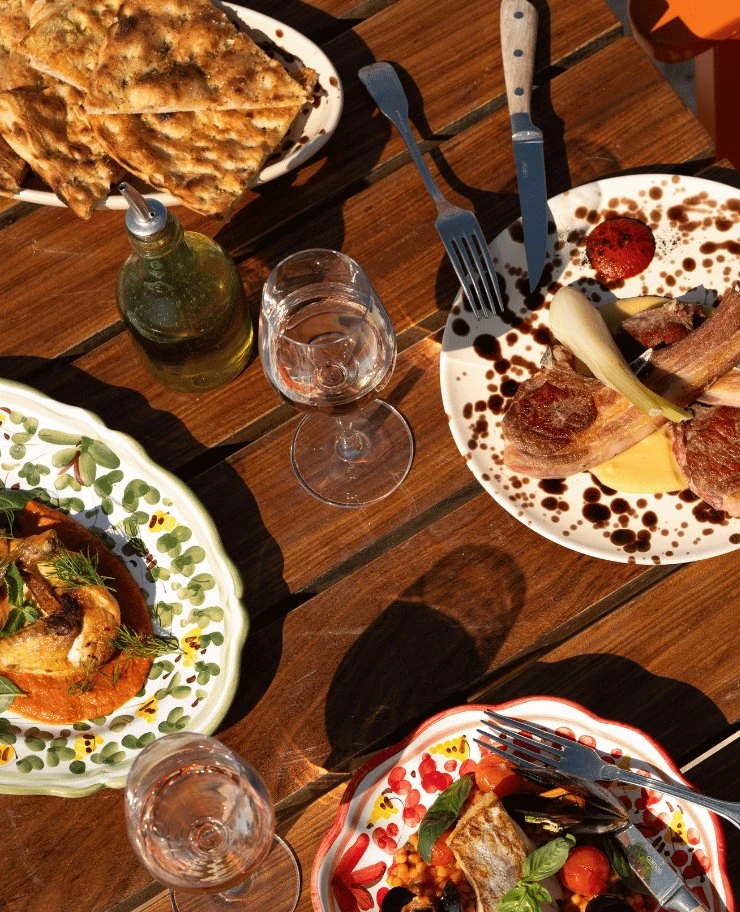 Table with plates of grilled meat, fish, bread, olive oil bottle, and two wine glasses on wood surface.