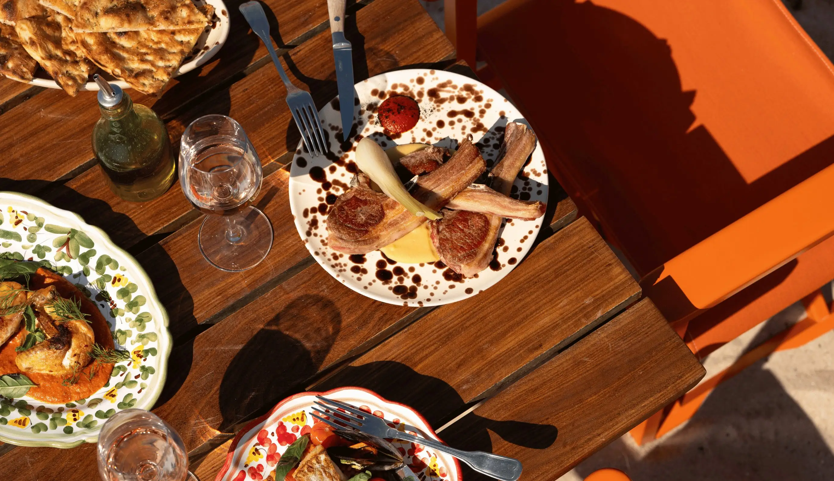 Table with three plates of gourmet food, two glasses of water, olive oil bottle, and orange chair nearby.