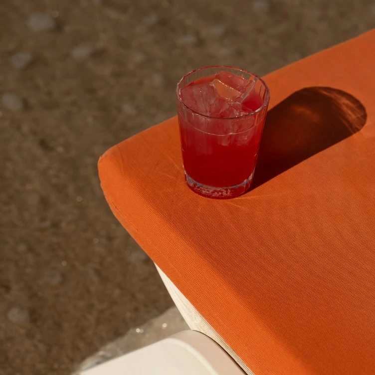 Red drink with ice cubes in a glass on an orange fabric surface casting a shadow.