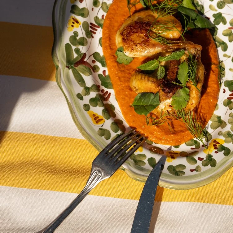 Roasted chicken on red sauce garnished with herbs, served on a floral plate with fork and knife.