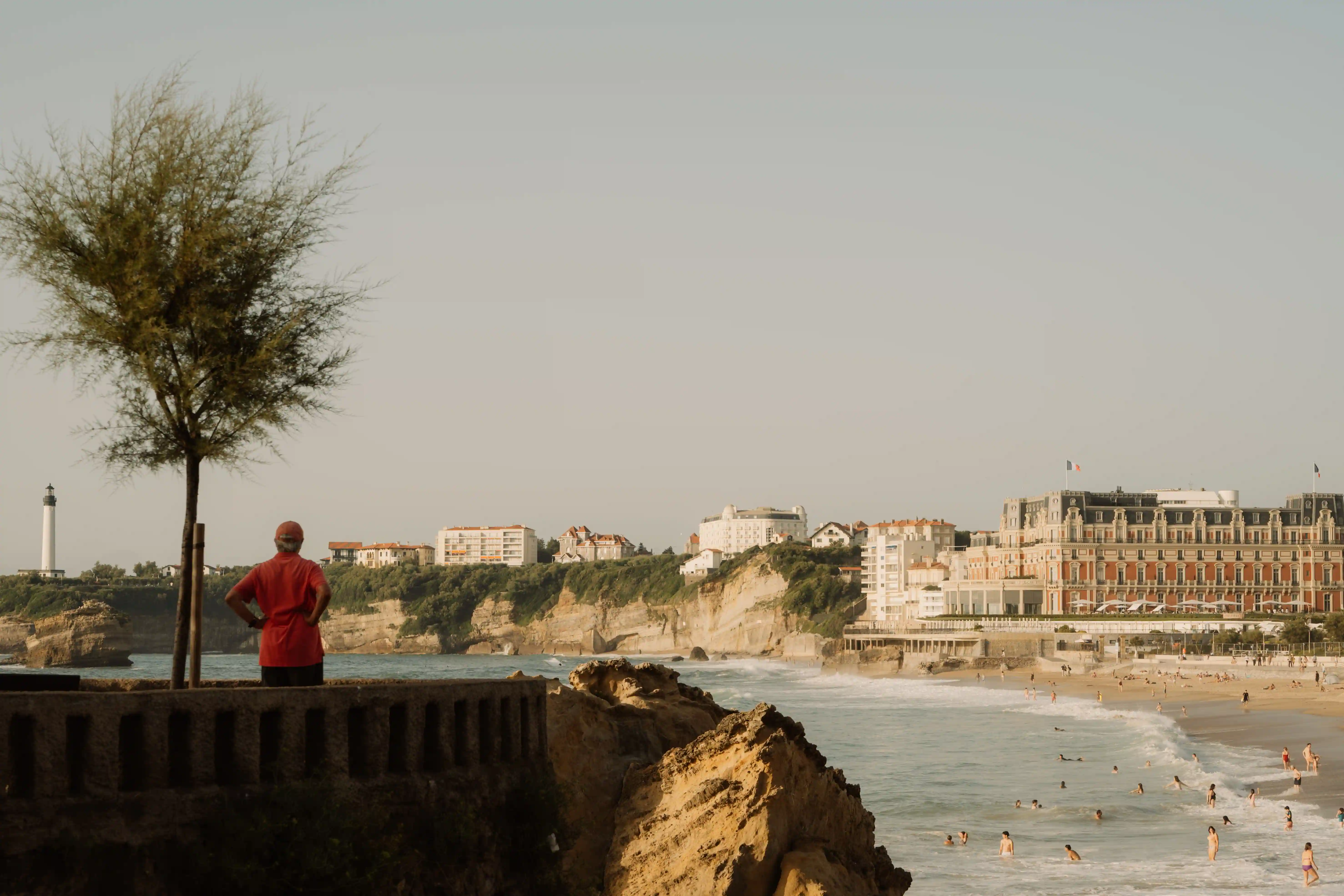 Person in red shirt standing by tree overlooking beach with swimmers and seaside buildings.