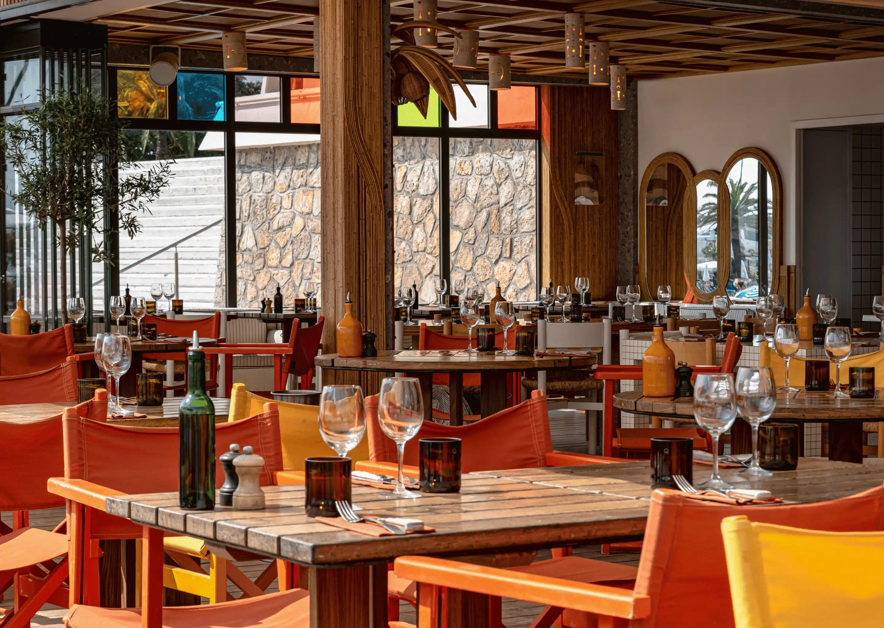 Empty restaurant dining area with wooden tables, colorful chairs, glassware, and stone wall windows.