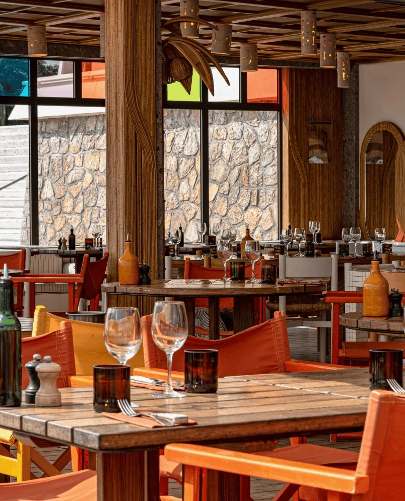 Indoor restaurant with wooden tables, orange chairs, glassware, and stone wall visible through windows.