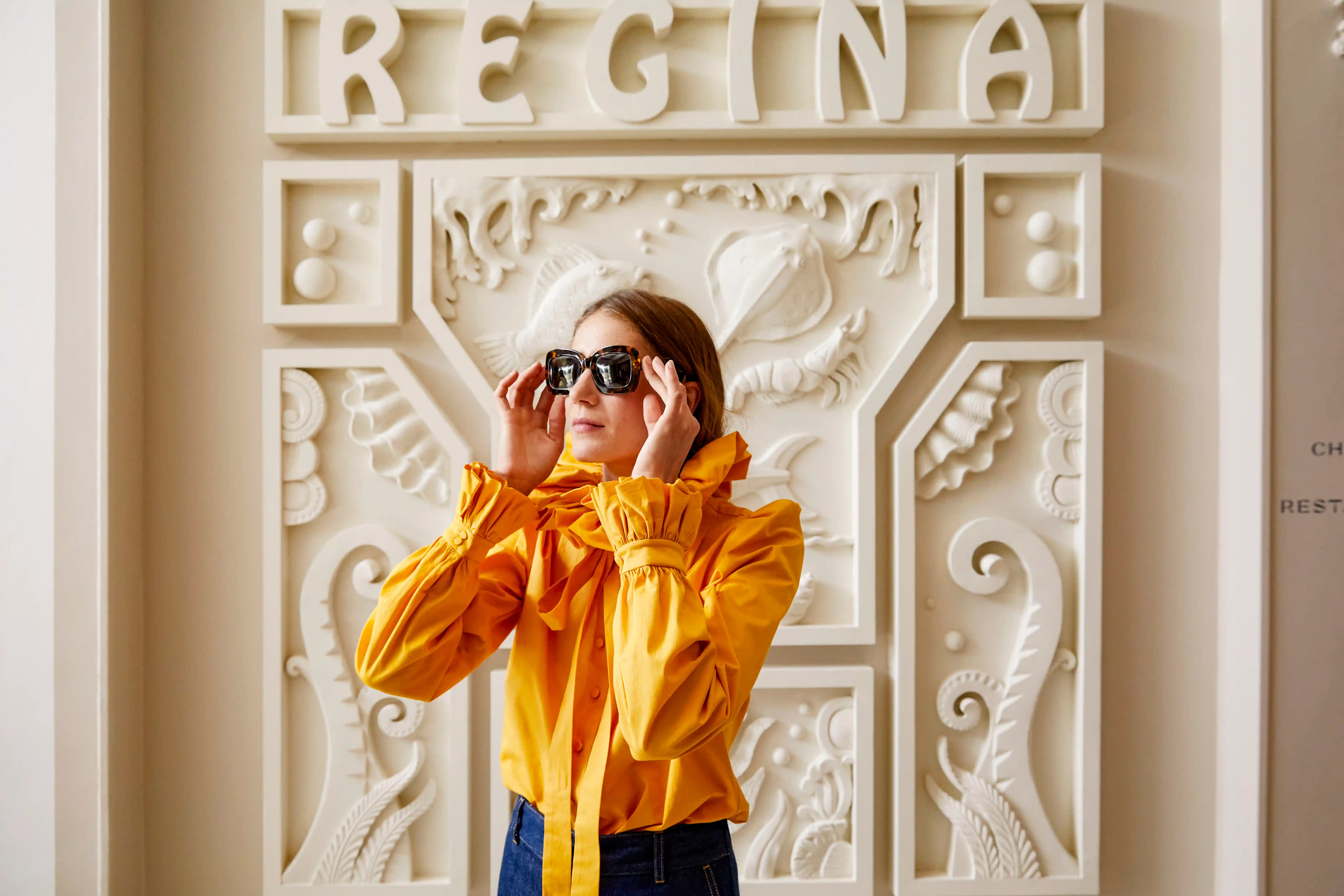 Woman in yellow blouse adjusting sunglasses in front of a decorative wall with 'REGINA' text.