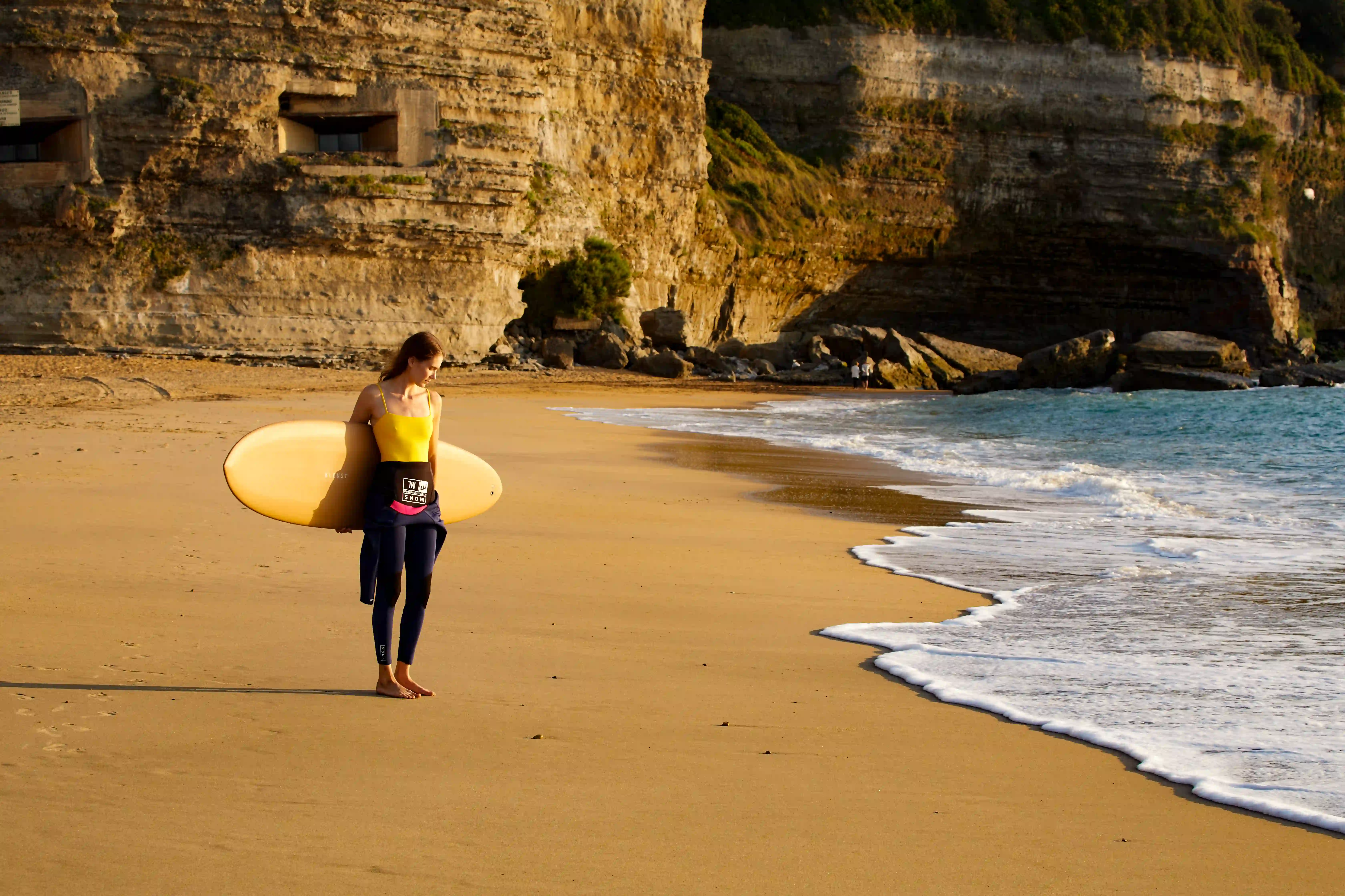 Woman in a yellow top holding a surfboard walking barefoot on a sandy beach near cliffs and ocean.