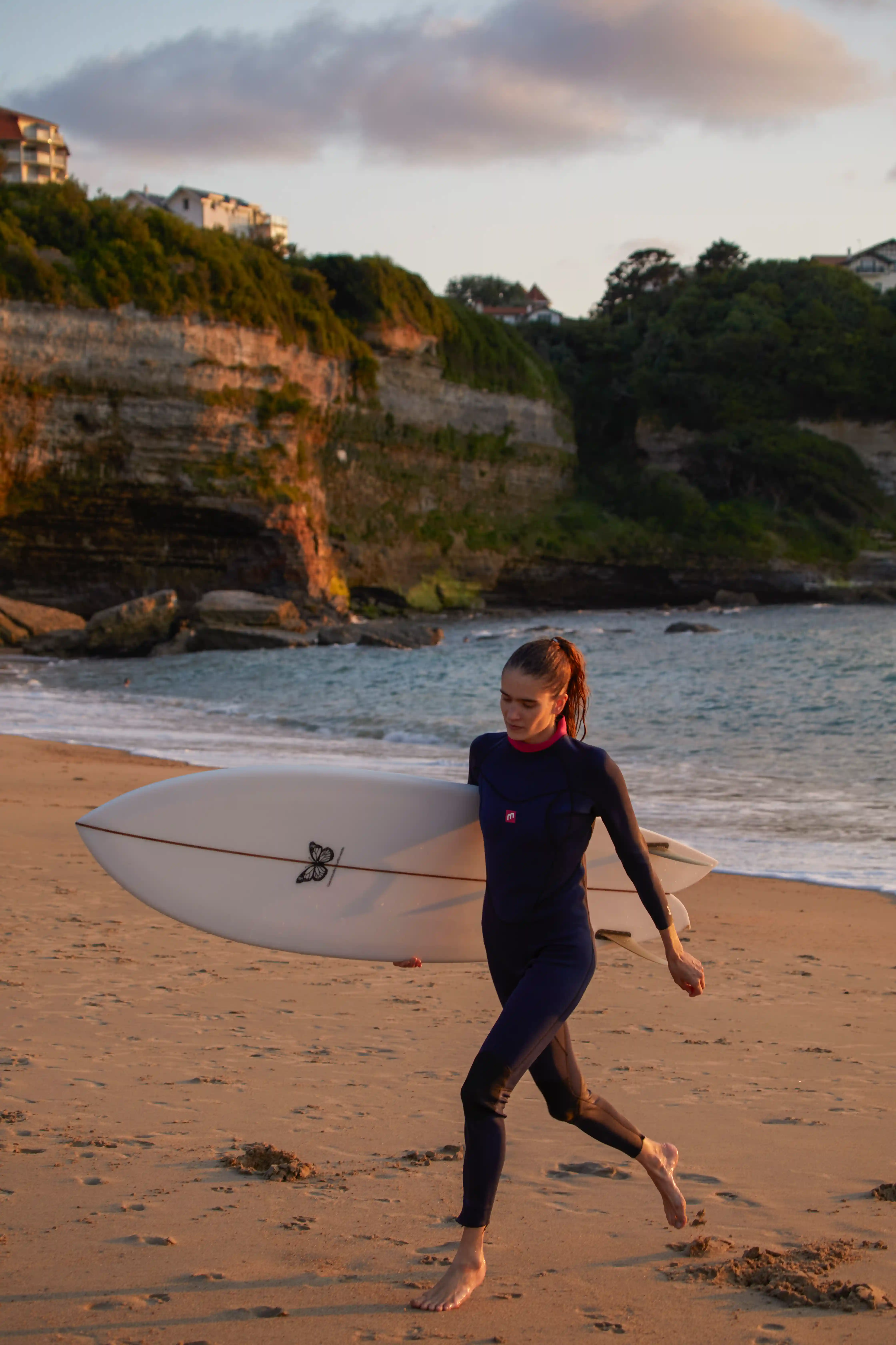 Woman in a wetsuit carrying a surfboard walking barefoot on a sandy beach near cliffs at sunset.