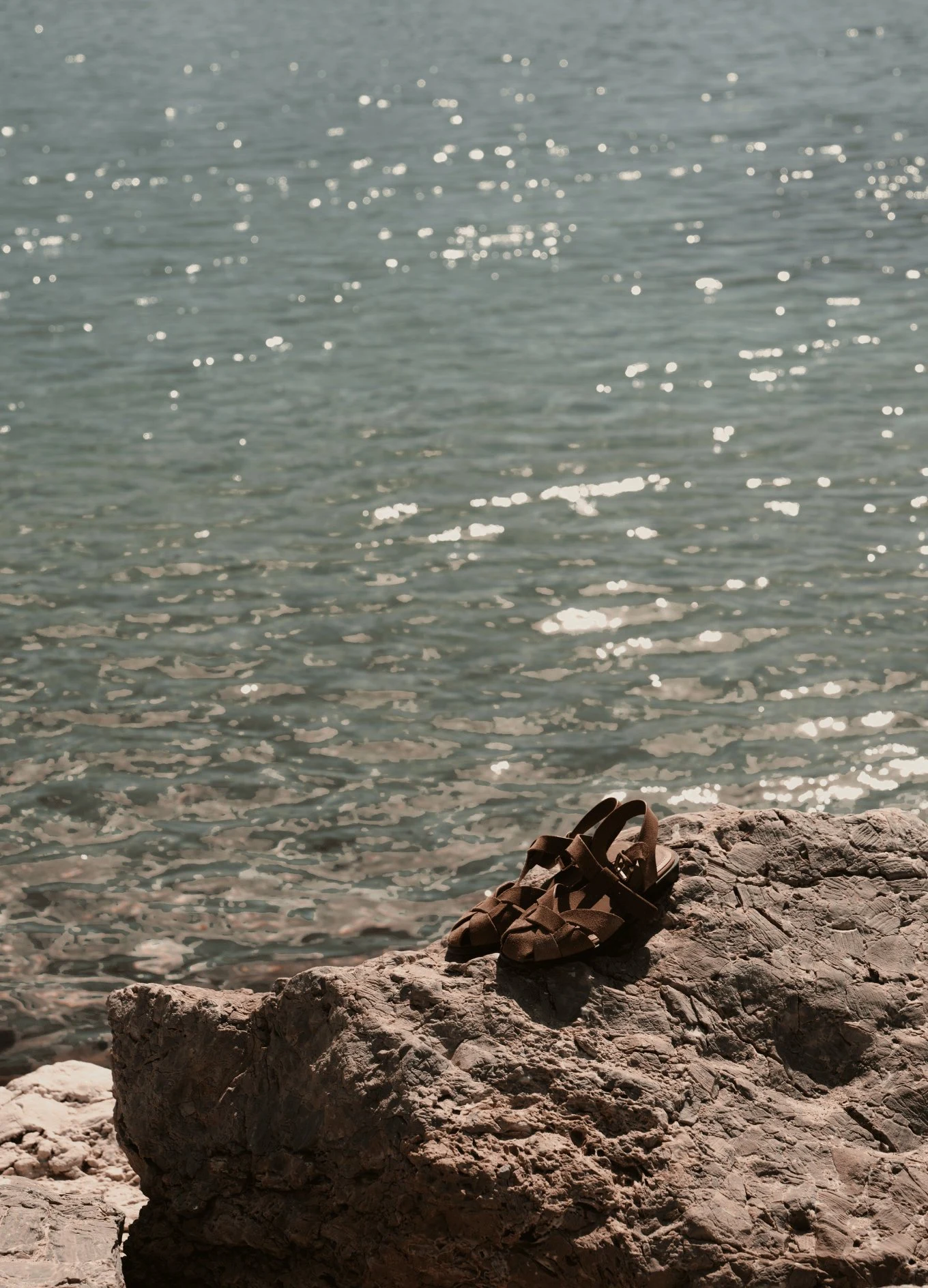 Brown woven sandals placed on a rock by the sparkling sea water at the beach.