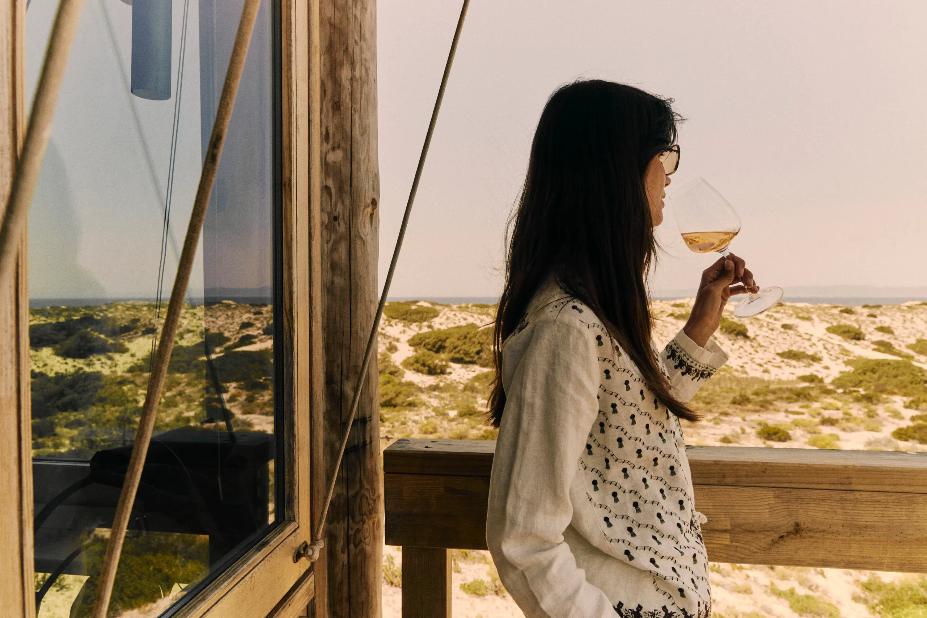 Woman with long dark hair and glasses drinking wine on a wooden balcony overlooking a sandy landscape.
