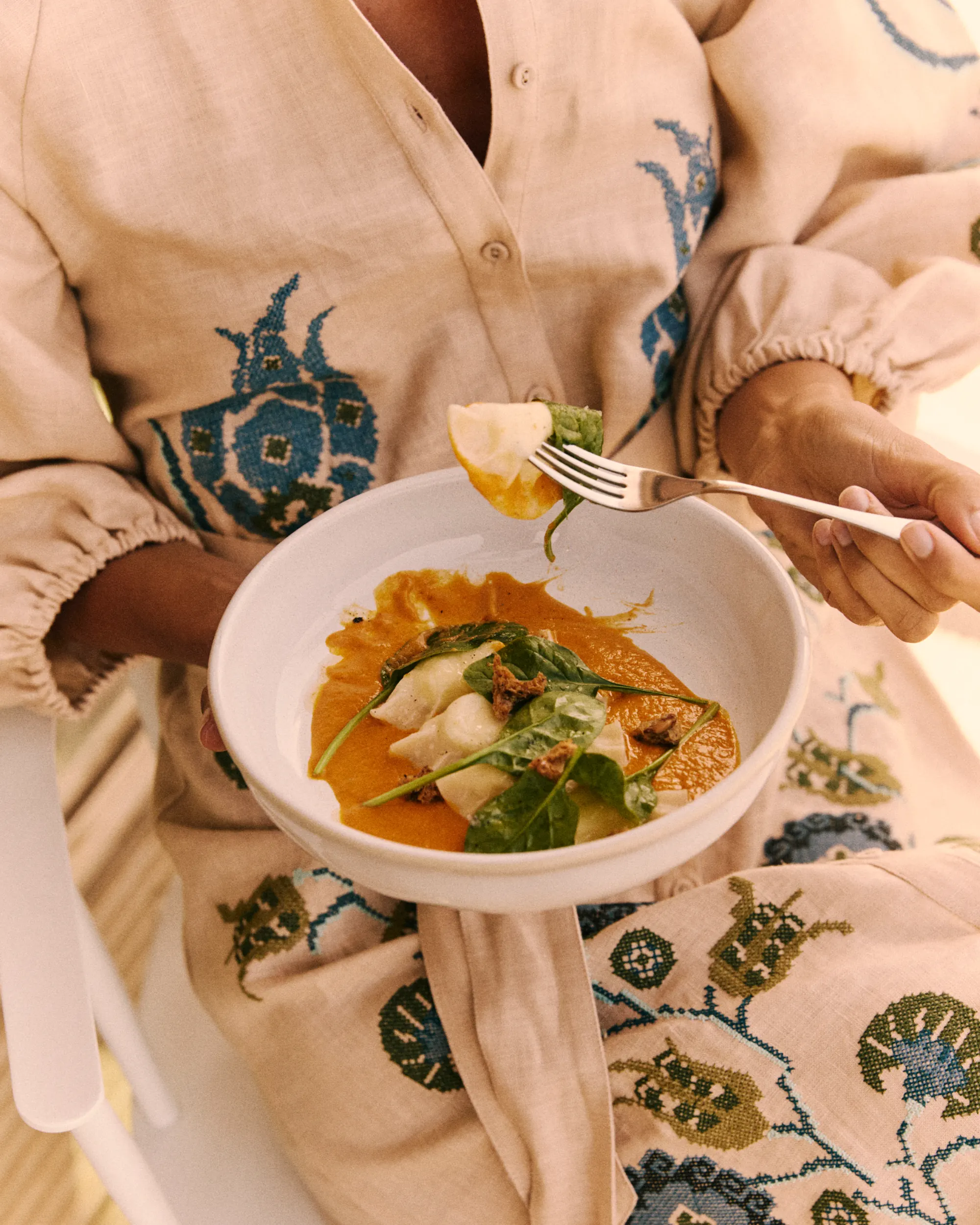 Person holding a bowl of ravioli with spinach and orange sauce, eating a piece with a fork.