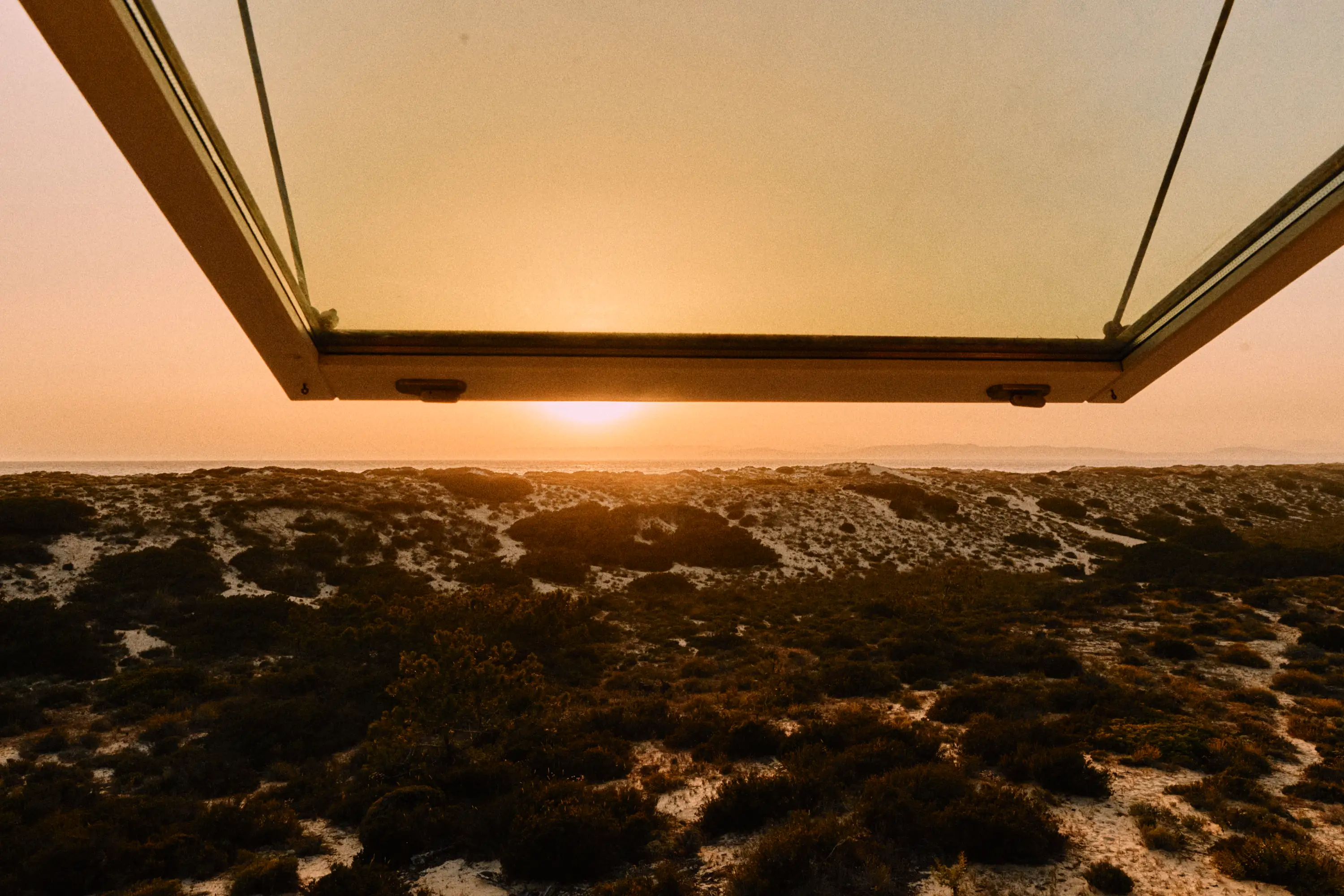 Sunset over sandy dunes with vegetation viewed through an open window at dusk.