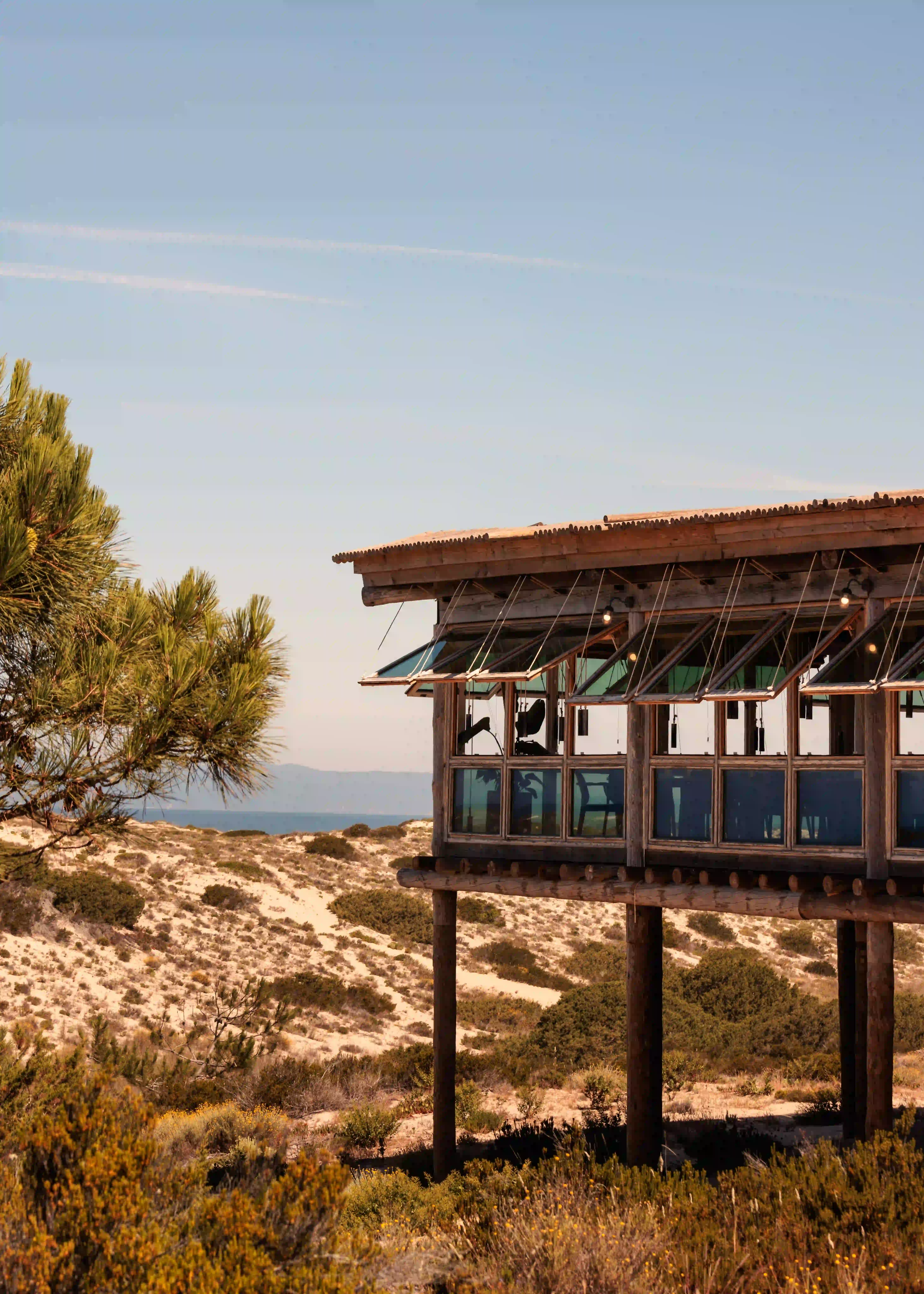 Wooden beachside building on stilts overlooking sandy dunes and bushes under a clear blue sky.