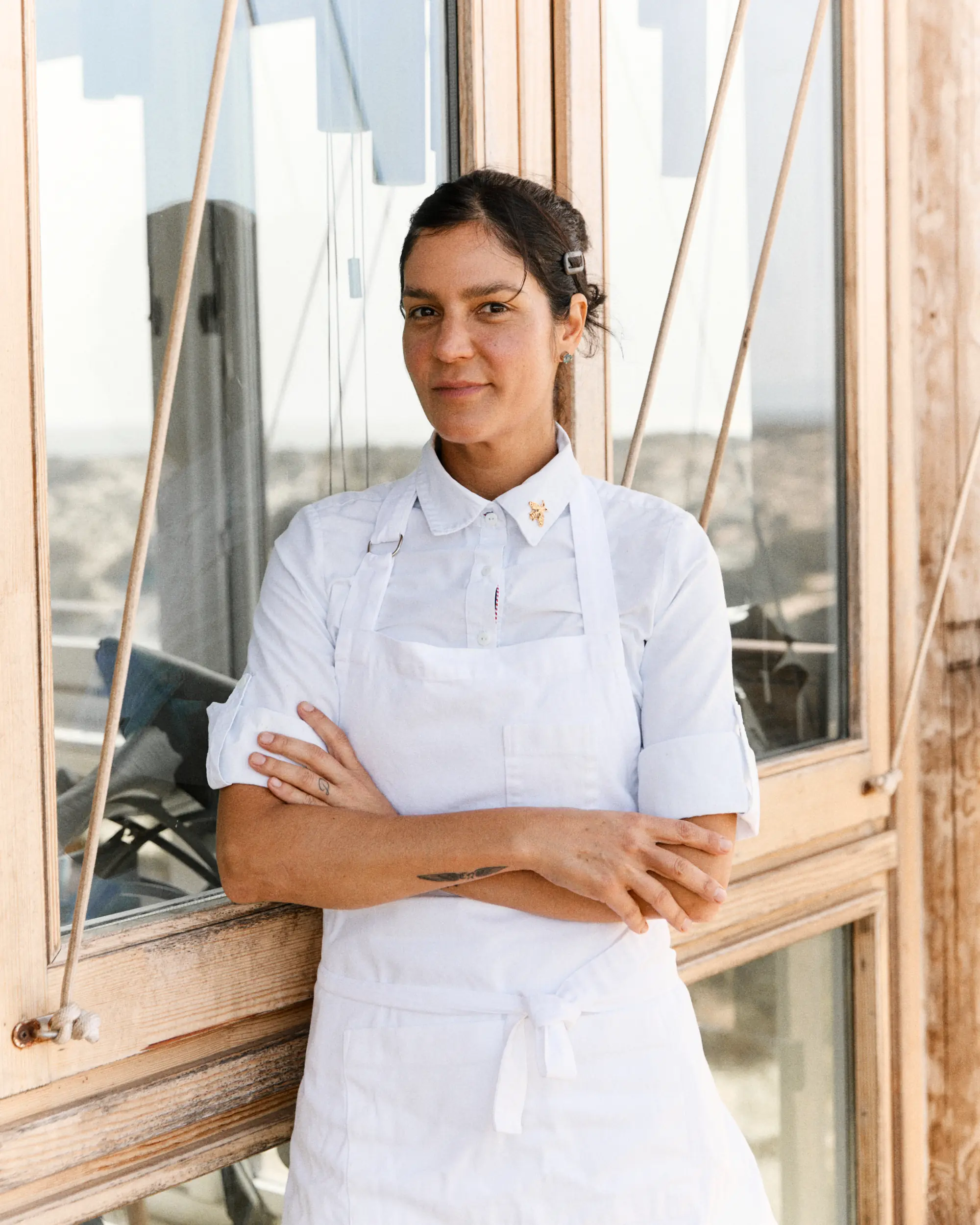 Woman with dark hair in white shirt and apron standing with arms crossed by wooden window.