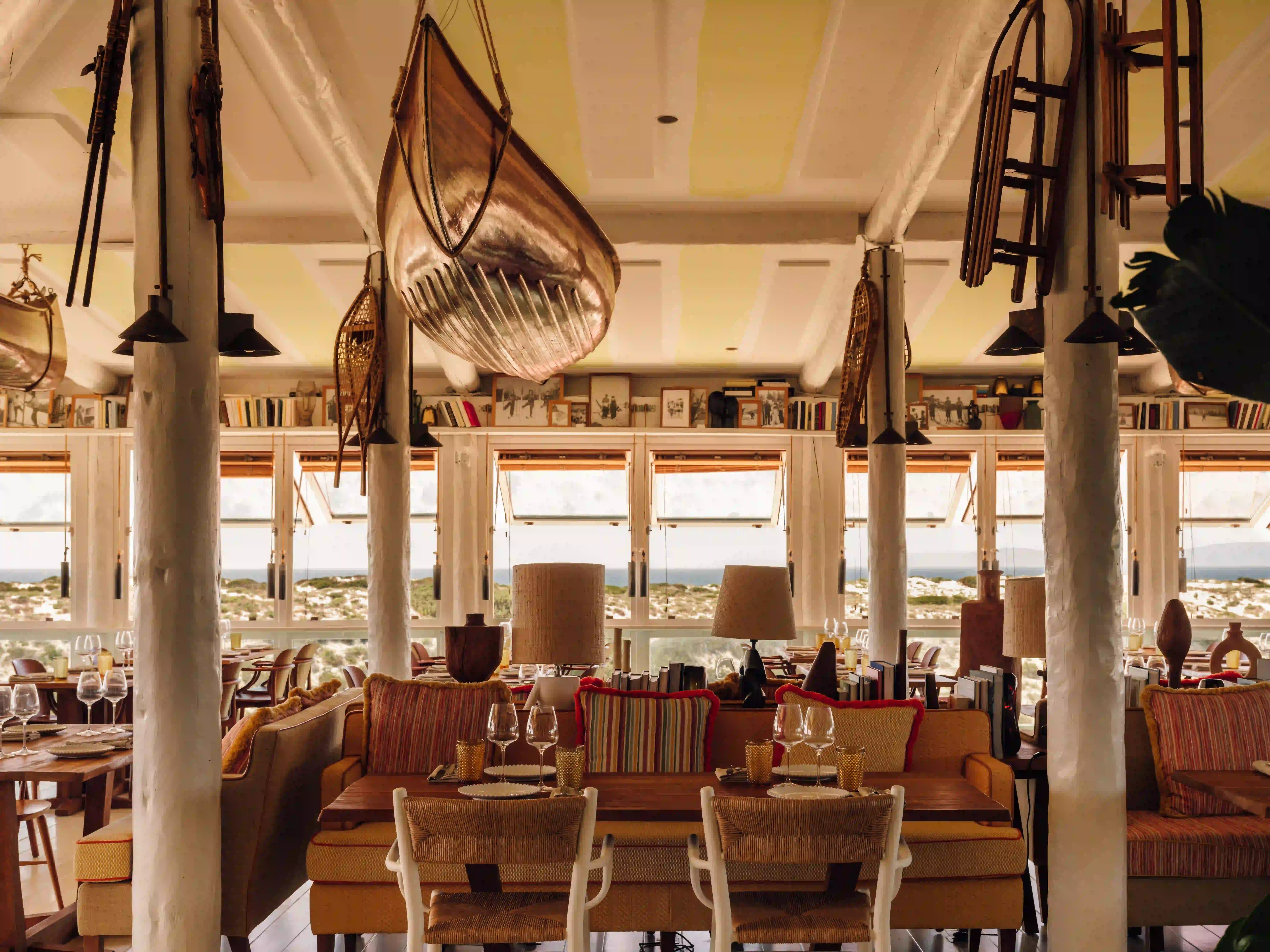 Cozy restaurant interior with wooden tables, striped cushions, hanging boat, and large windows with dune views.