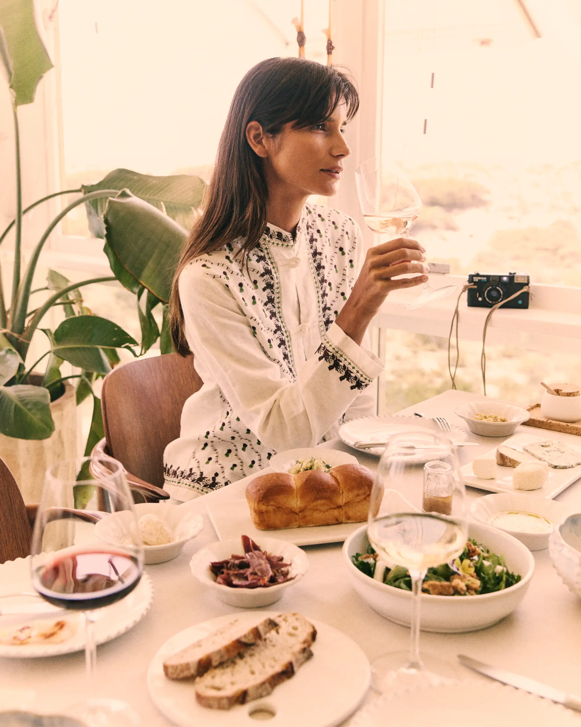 Woman in white embroidered top holding a glass of white wine at a table with bread and salad.