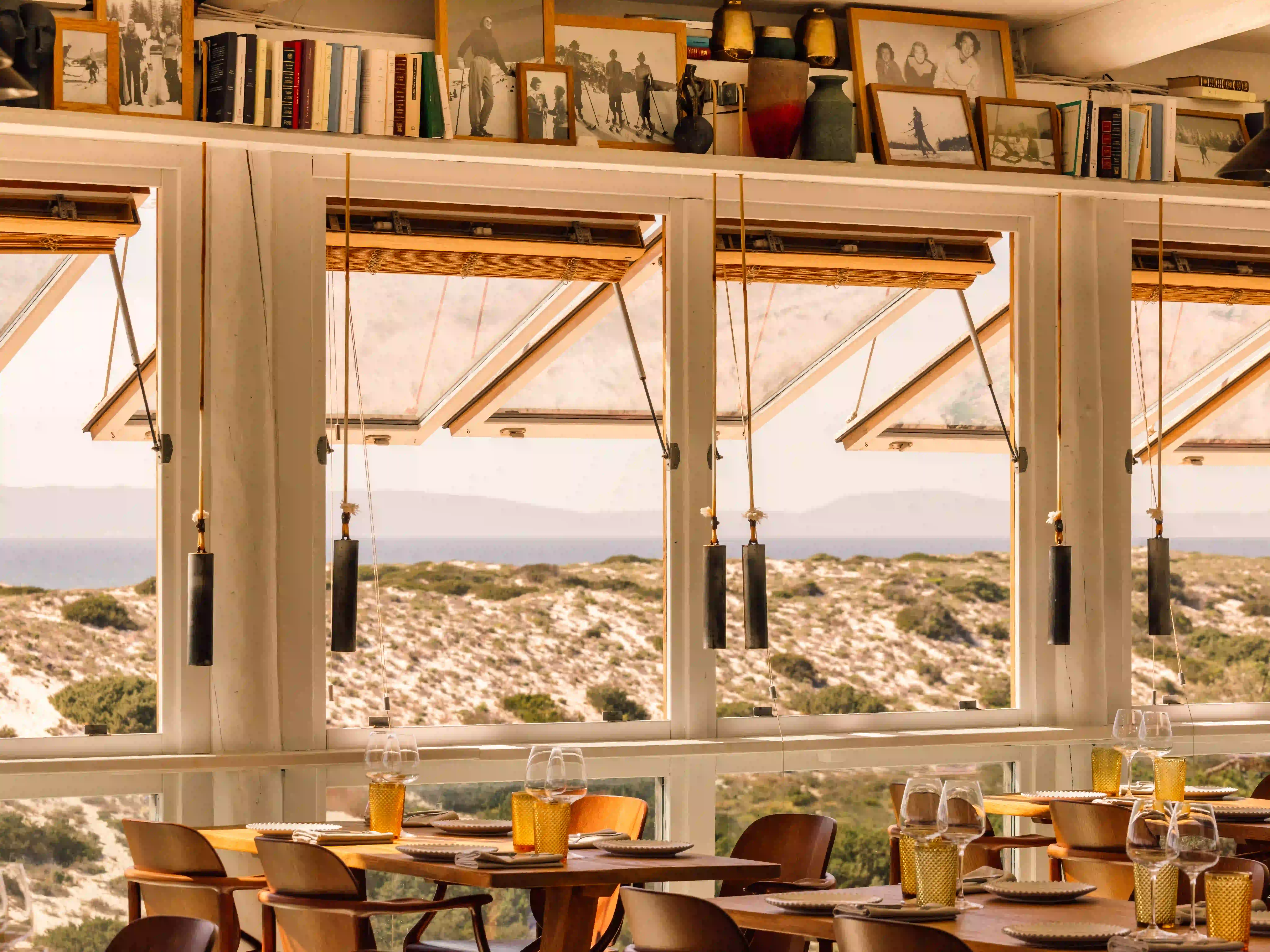 Cozy dining area with wooden tables, chairs, and large windows overlooking sandy dunes and sea.