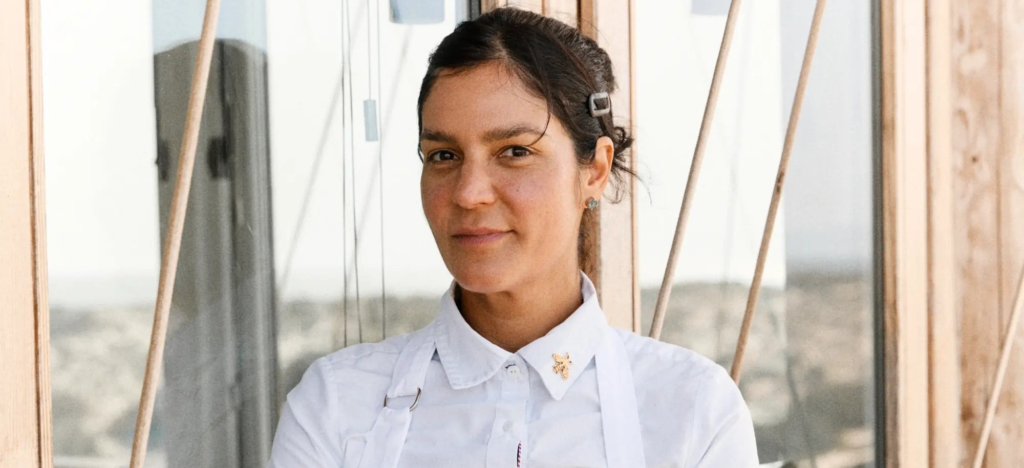 Woman with dark hair in a white shirt and apron stands in front of large glass windows and wooden frames.