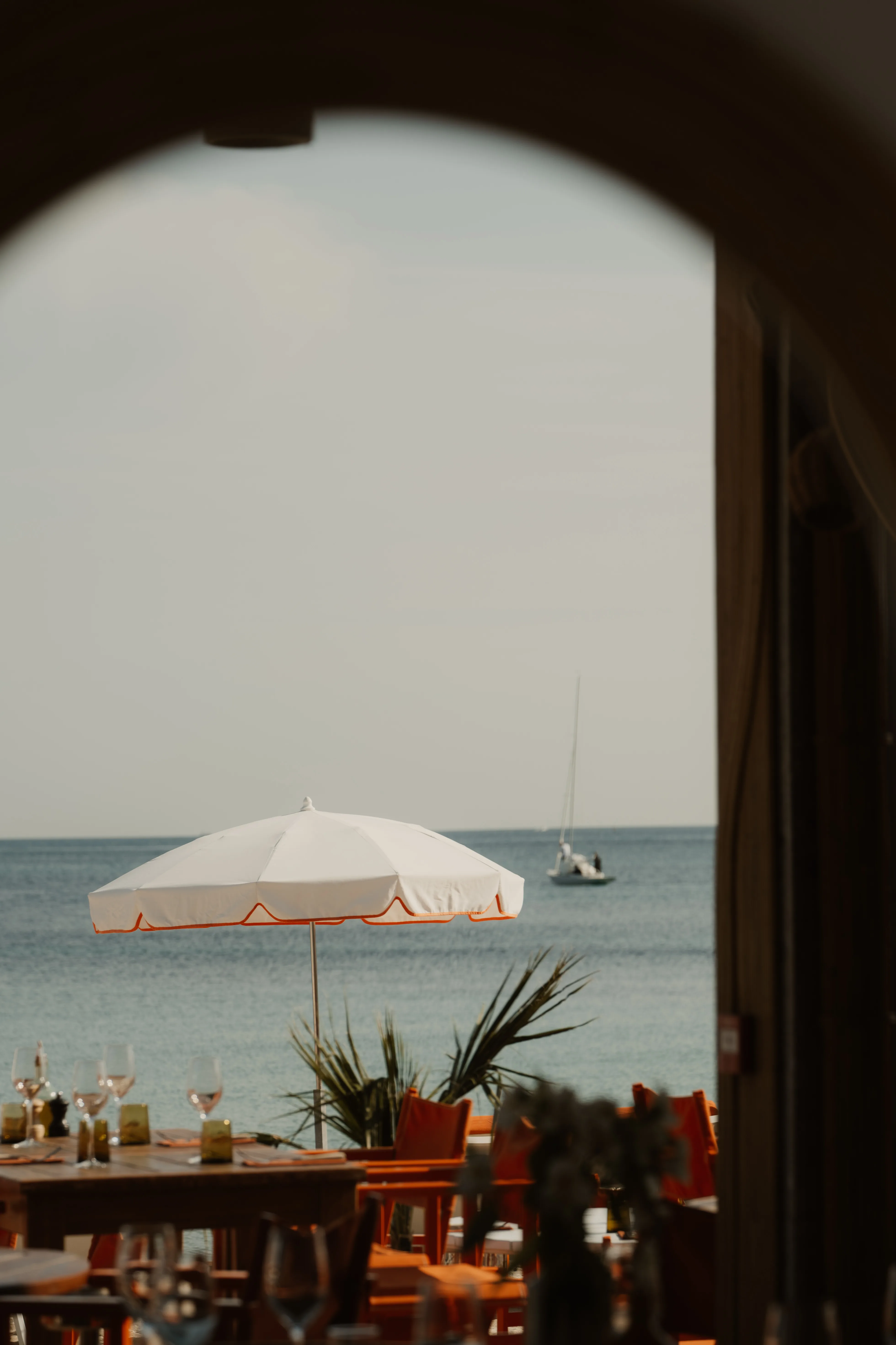 Beachside dining area with a white umbrella, orange chairs, and a sailboat on the blue sea.