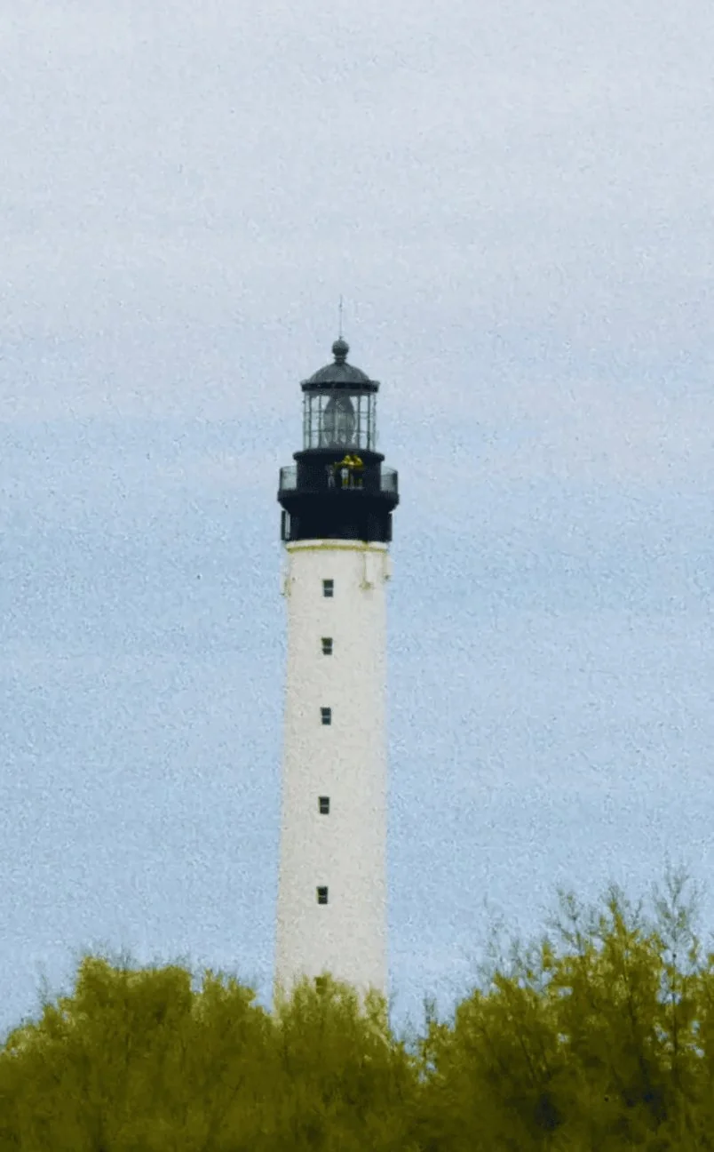 Tall white lighthouse with black top above green trees against a blue sky.