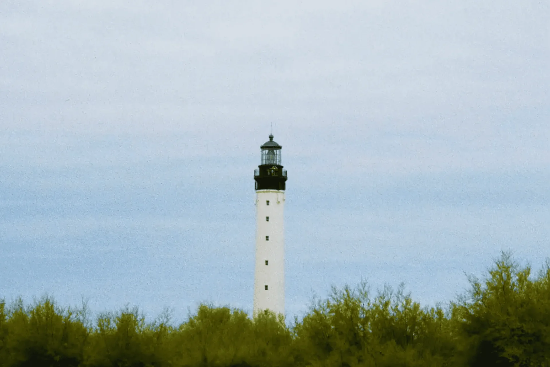 Tall white lighthouse with black top above green trees under a cloudy sky.