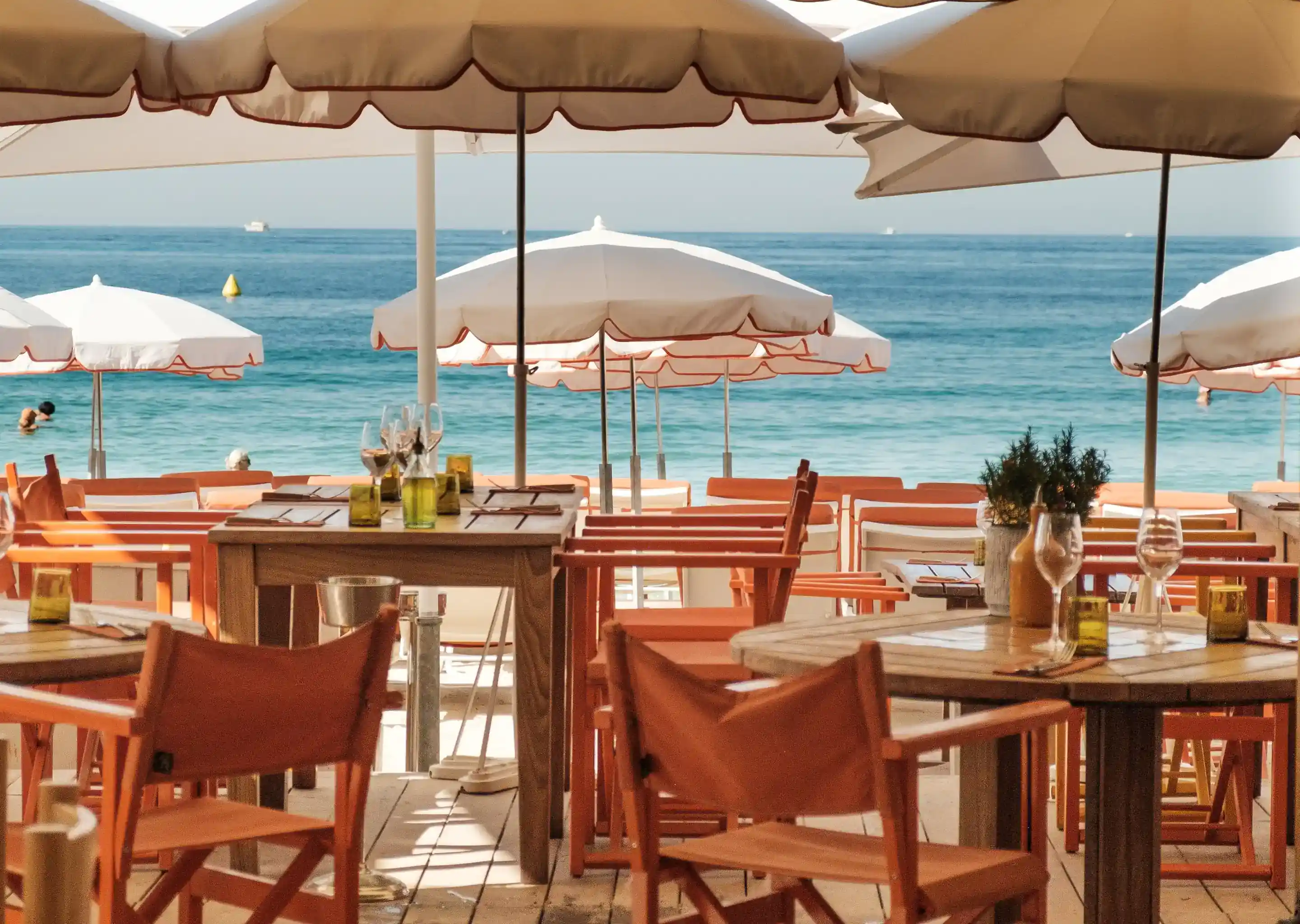 Beachside restaurant with wooden tables and orange chairs under umbrellas overlooking a calm sea.