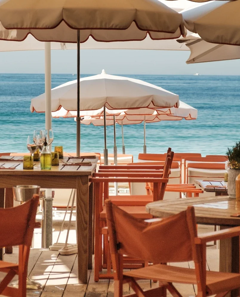 Outdoor beachside dining with orange chairs, tables, white umbrellas, and ocean view.
