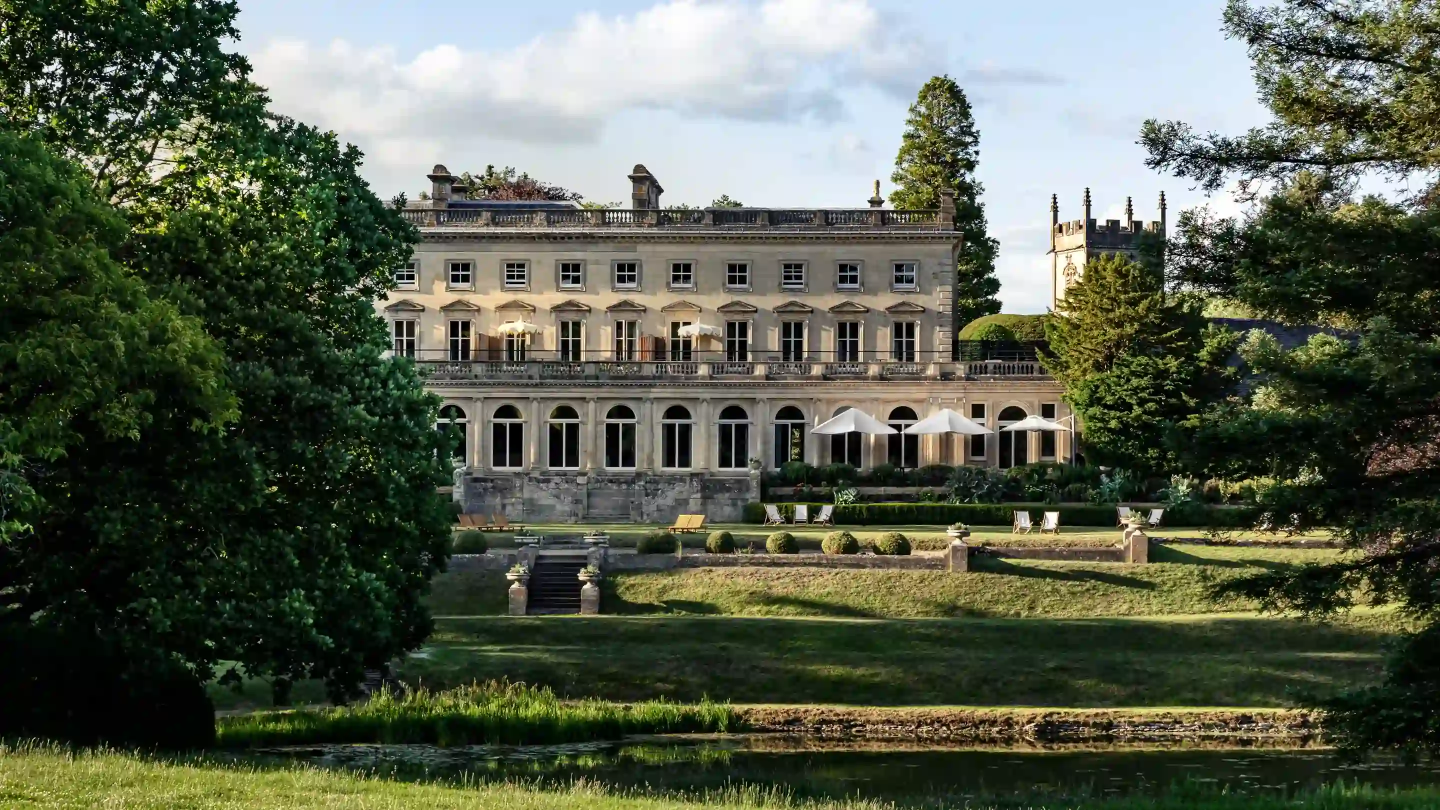 Large stone manor house with tall windows, umbrellas, surrounded by lush trees and a pond in front.