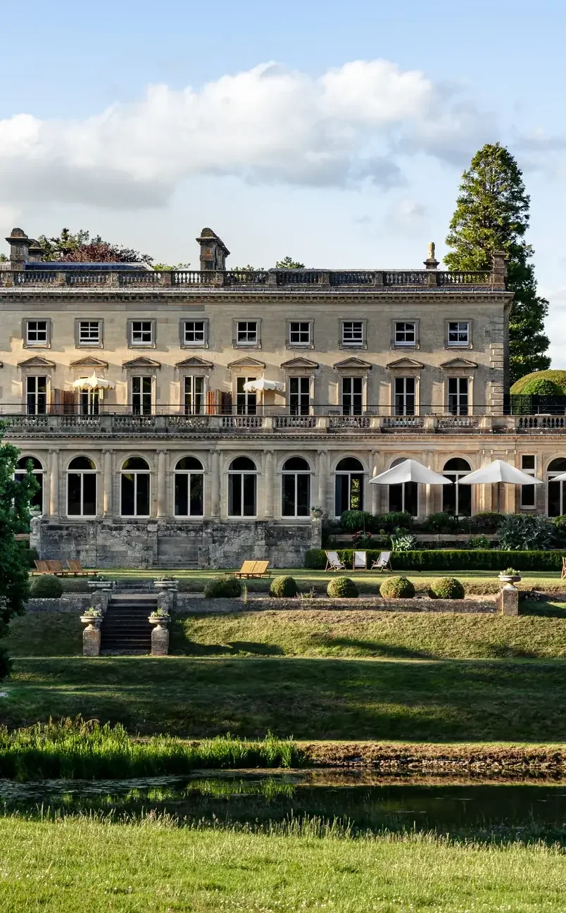 Large historic stone mansion with arched windows, patio umbrellas, green lawn, and pond in front.
