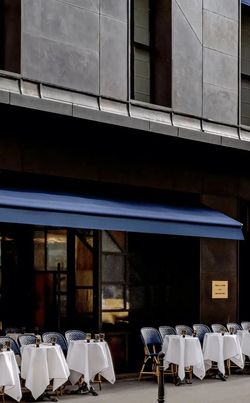 Outdoor cafe seating with white tablecloths and blue chairs under a blue awning on a city sidewalk.