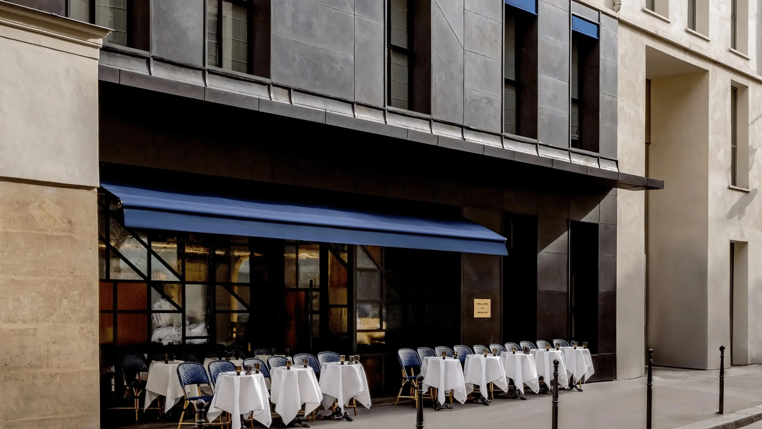 Outdoor café seating with blue chairs and white tablecloths under a blue awning along a city street.