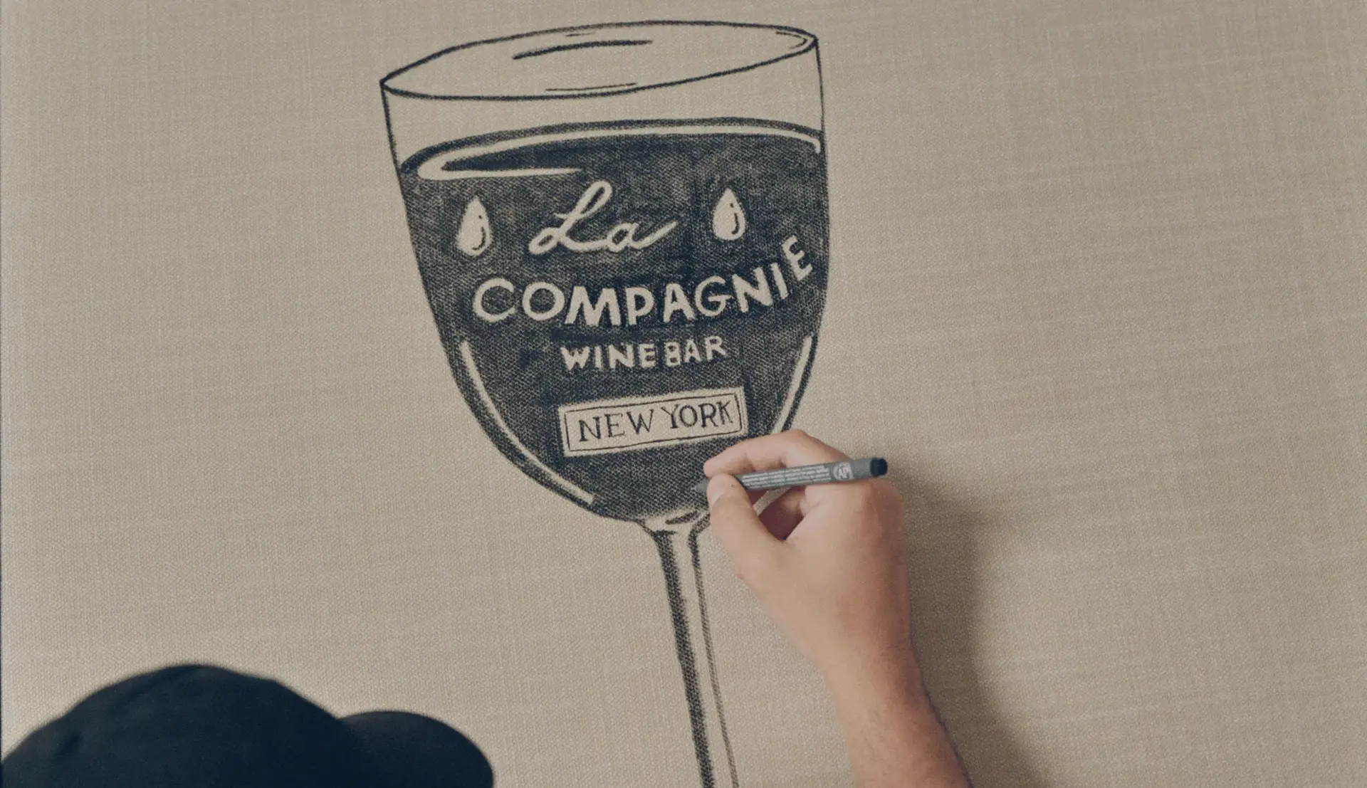 Hand drawing a wine glass with text 'La Compagnie Wine Bar New York' on a beige canvas.