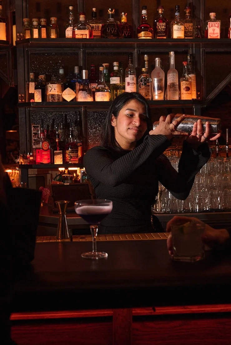 A bartender shaking a cocktail shaker behind a bar with bottles and glasses on shelves.